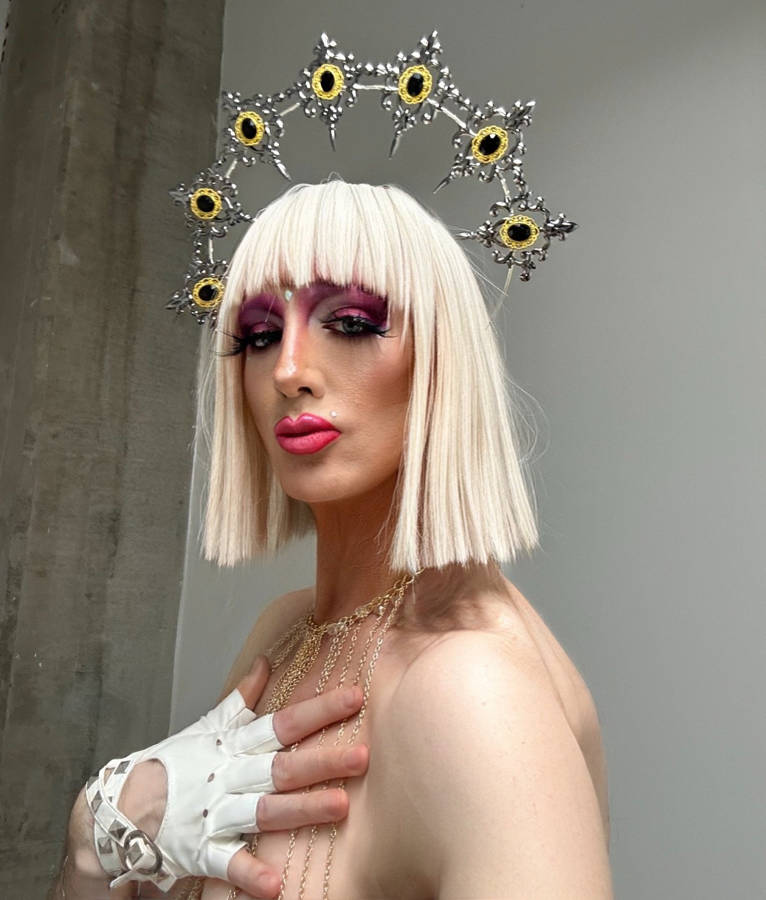 Person with platinum blonde bob haircut and bangs, wearing pink eyeshadow, bold pink lipstick, a decorative headpiece with black and yellow sunflowers, gold chains, and a white fingerless glove, posing with hand on chest.