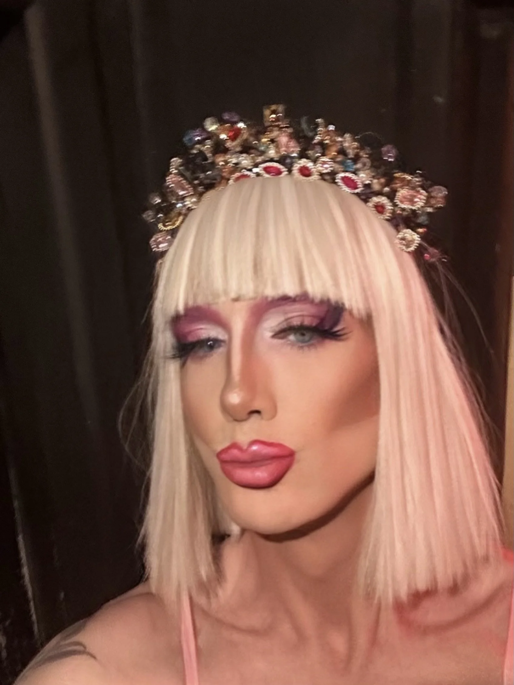 A woman with platinum blonde hair and blunt bangs, wearing a jeweled crown with red and black stones, and bold makeup with pink eyeshadow, thick false eyelashes, and glossy pink lipstick.