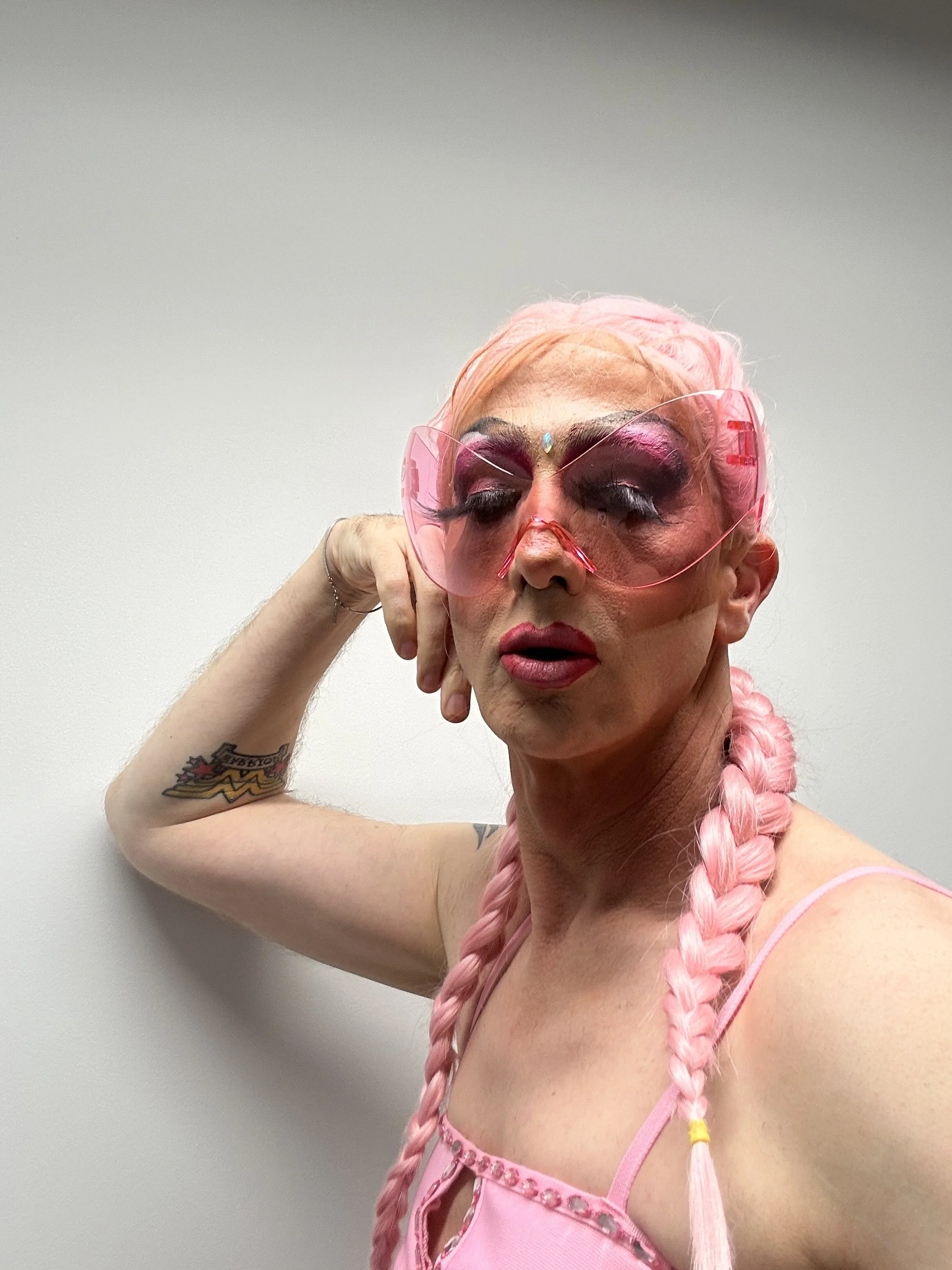 A person with long pink braided hair wearing pink sunglasses and pink makeup, leaning their head on their hand with a serious expression, standing against a plain white wall.