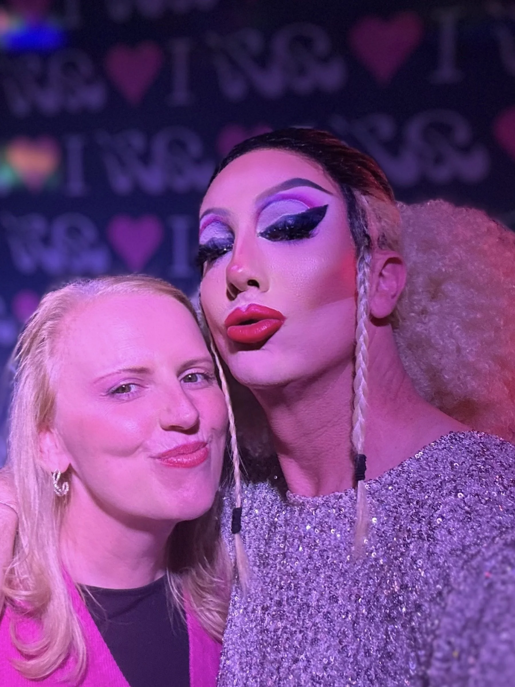Two individuals taking a close selfie in a setting with a stage background. One has blonde hair and wears a pink top, the other has dramatic makeup with bold eyeliner, red lipstick, and styled hair.