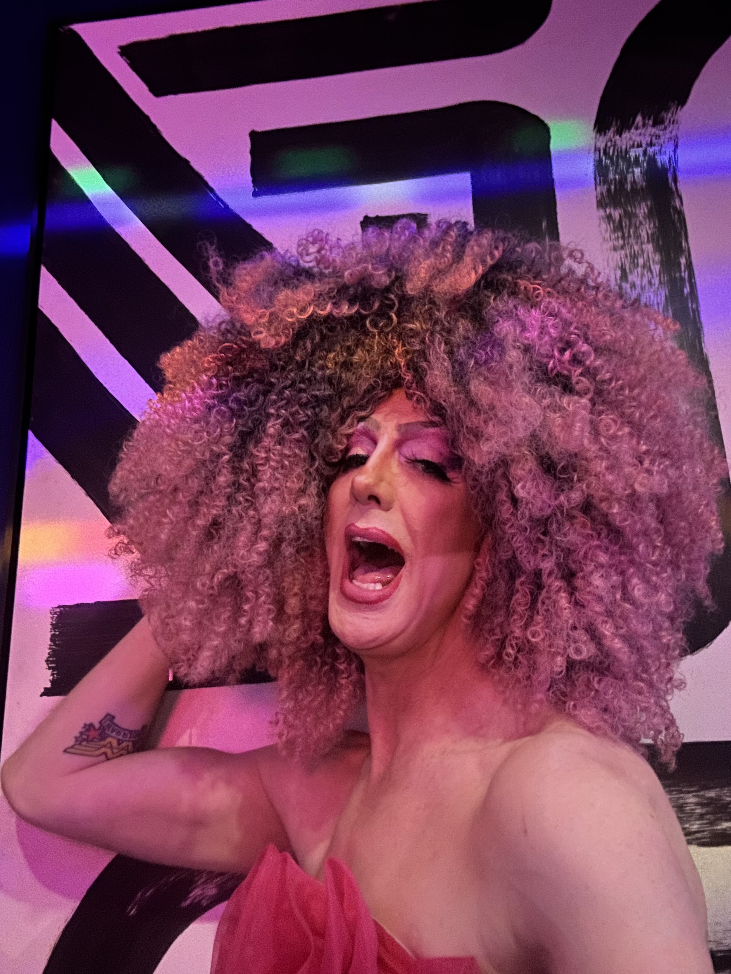 A person with curly blonde hair singing or shouting with their eyes closed and mouth open, wearing a pink strapless dress, against a black and white abstract geometric background.