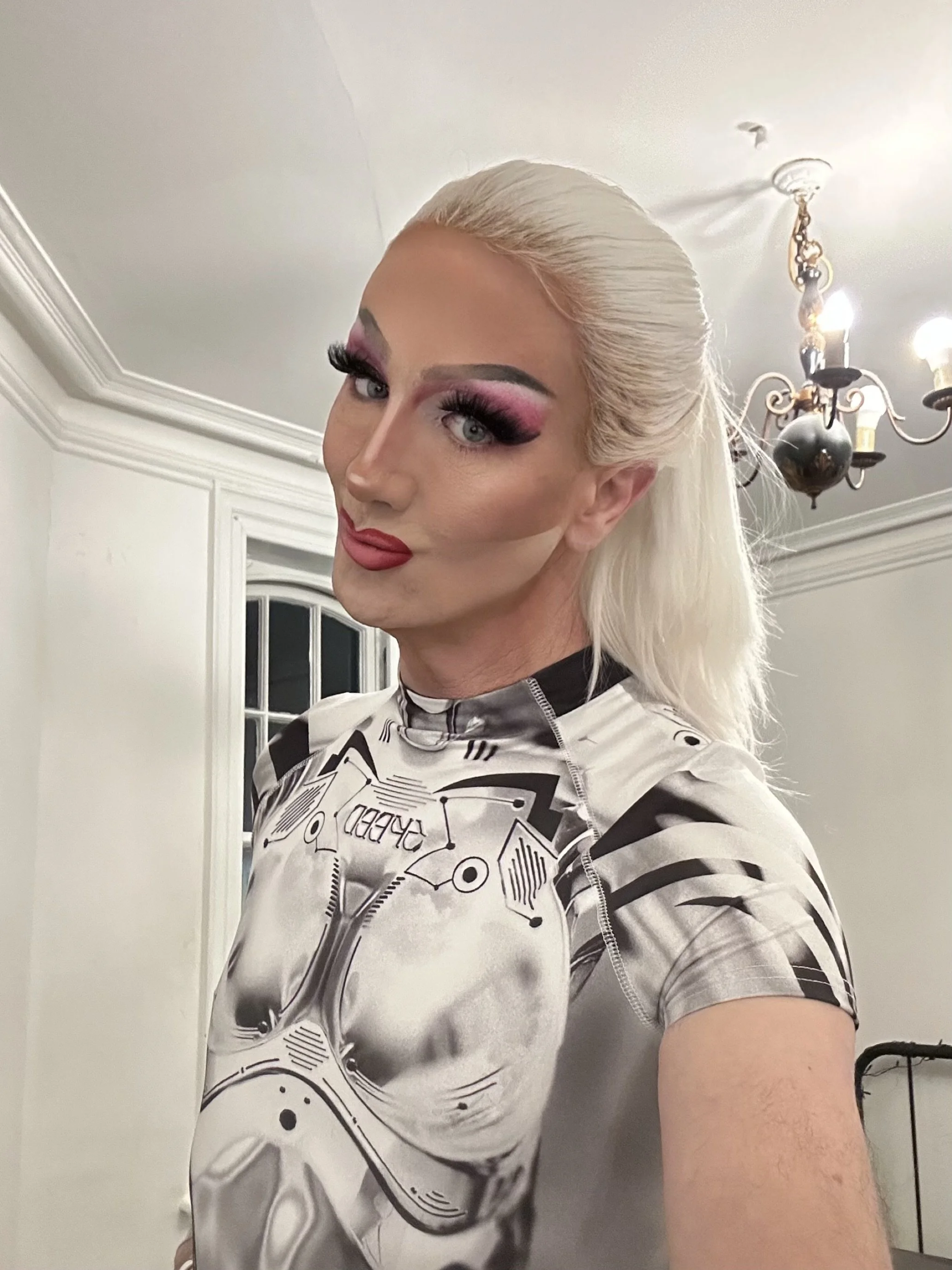 Person with platinum blonde hair styled back, wearing dramatic makeup with pink and black eyeshadow, false eyelashes, and red lipstick. They are dressed in a form-fitting shirt with a black and white graphic print of a robot or mechanical design. The