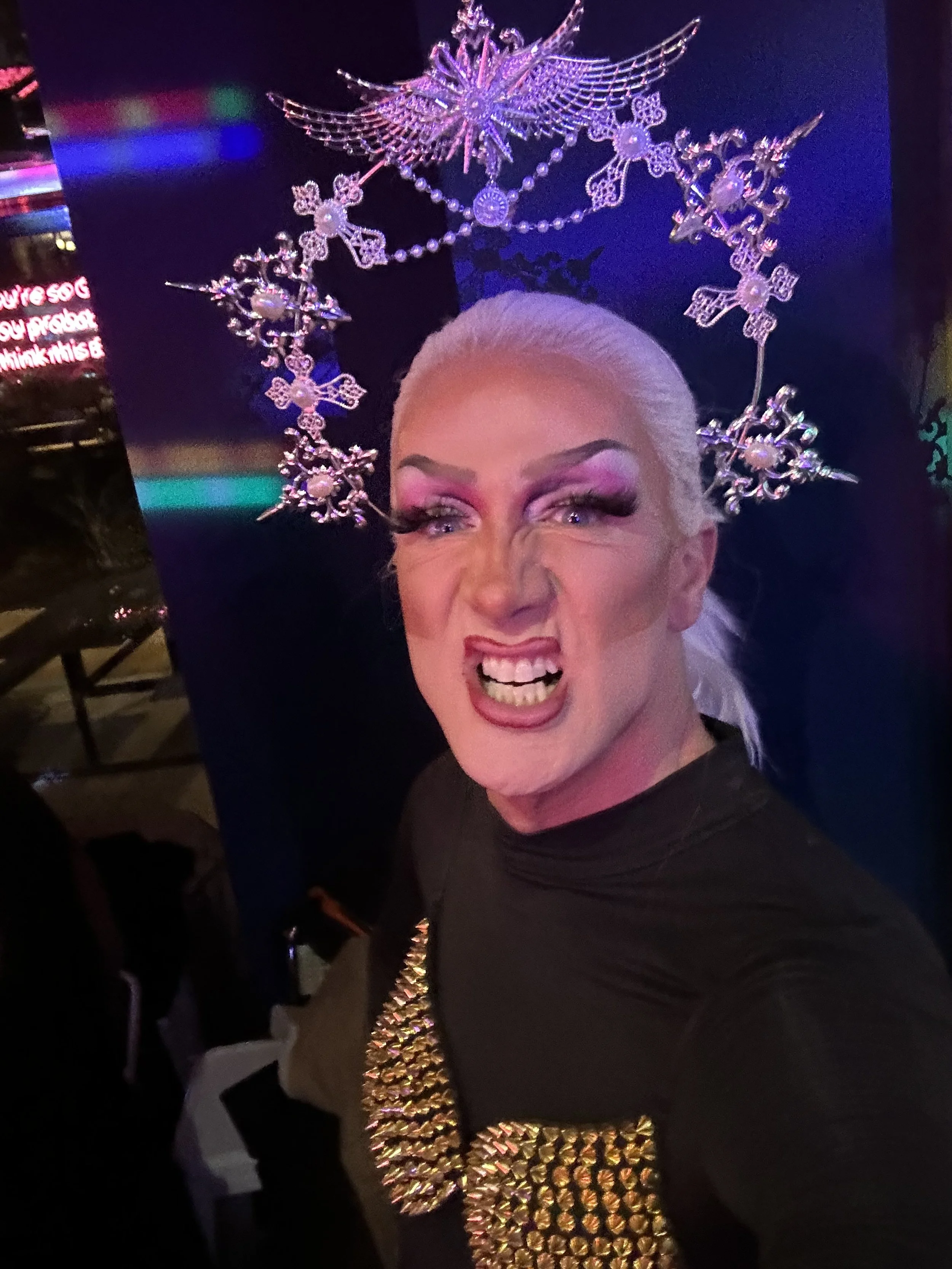 Person with platinum blonde hair wearing a large, decorated necklace and an elaborate crown with star, snowflake, and angel designs, posing with a fierce expression in a dark, colorful environment.