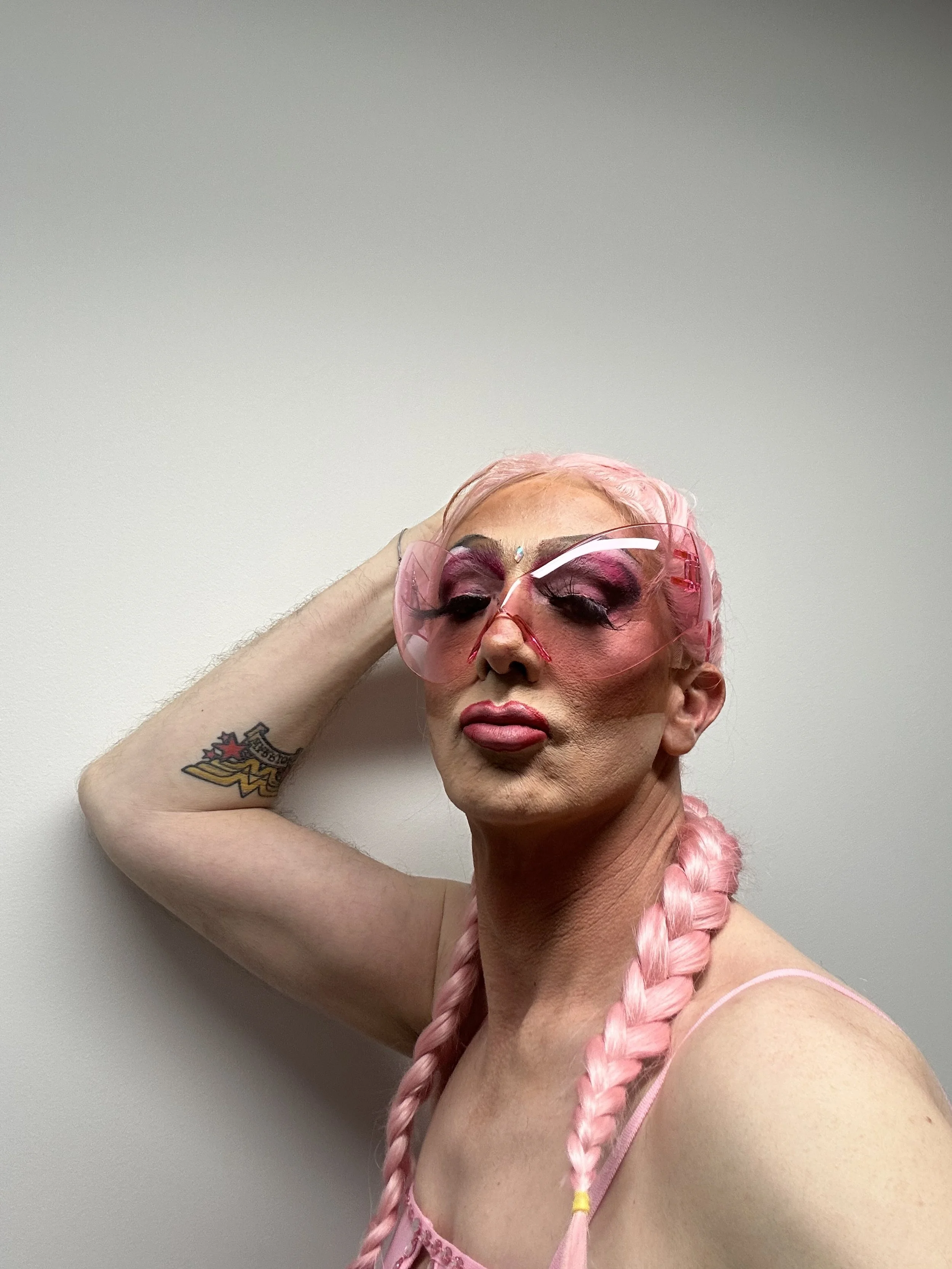 Person with pink braided hair, wearing pink sunglasses, pink makeup, and a pink top, posing against a plain white wall.