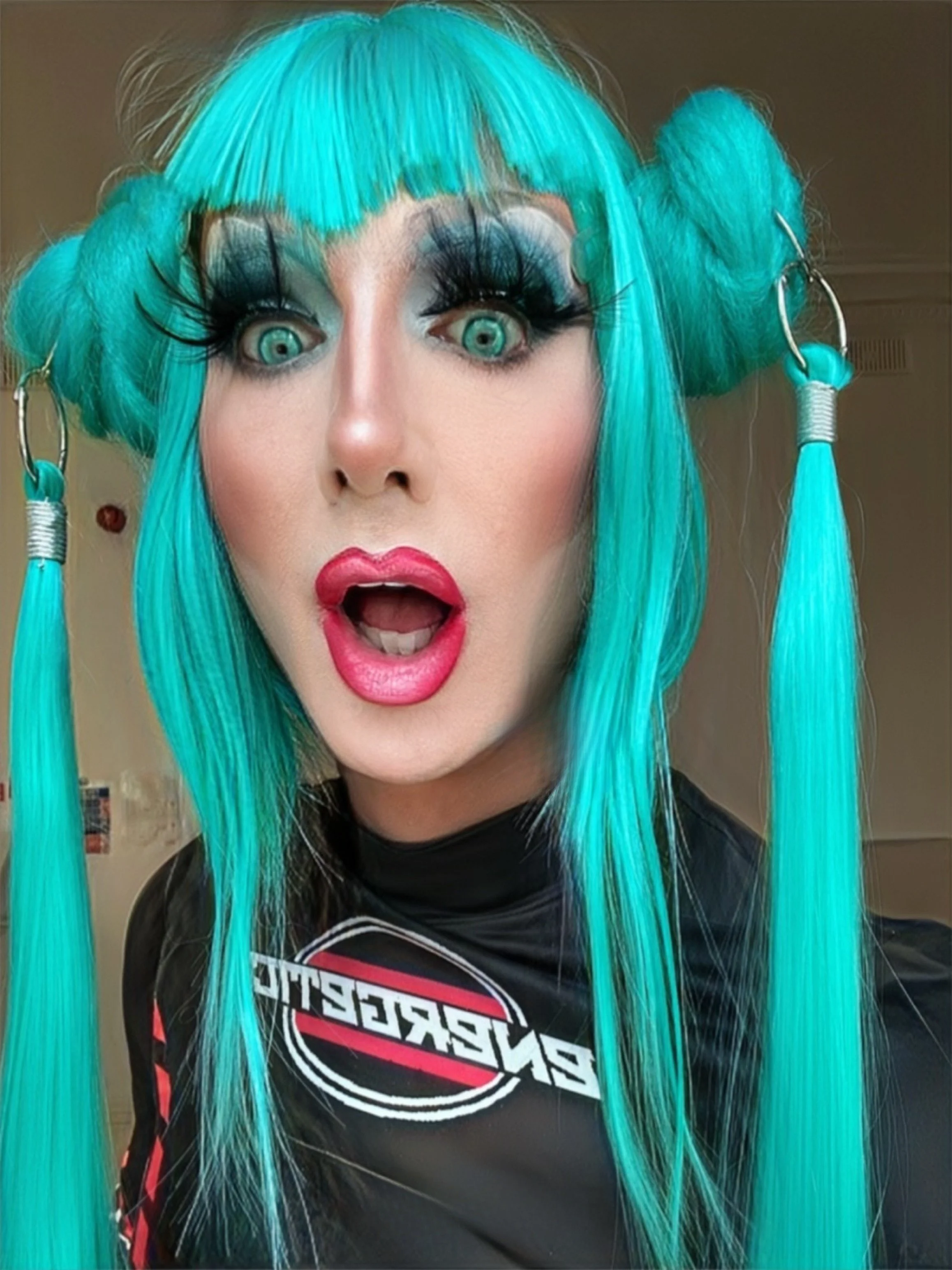 Person with turquoise hair styled in pigtails, wearing bold makeup with wide eyes, dark eye shadow, and bright pink lipstick, dressed in a black shirt with a graphic design, surprised expression.