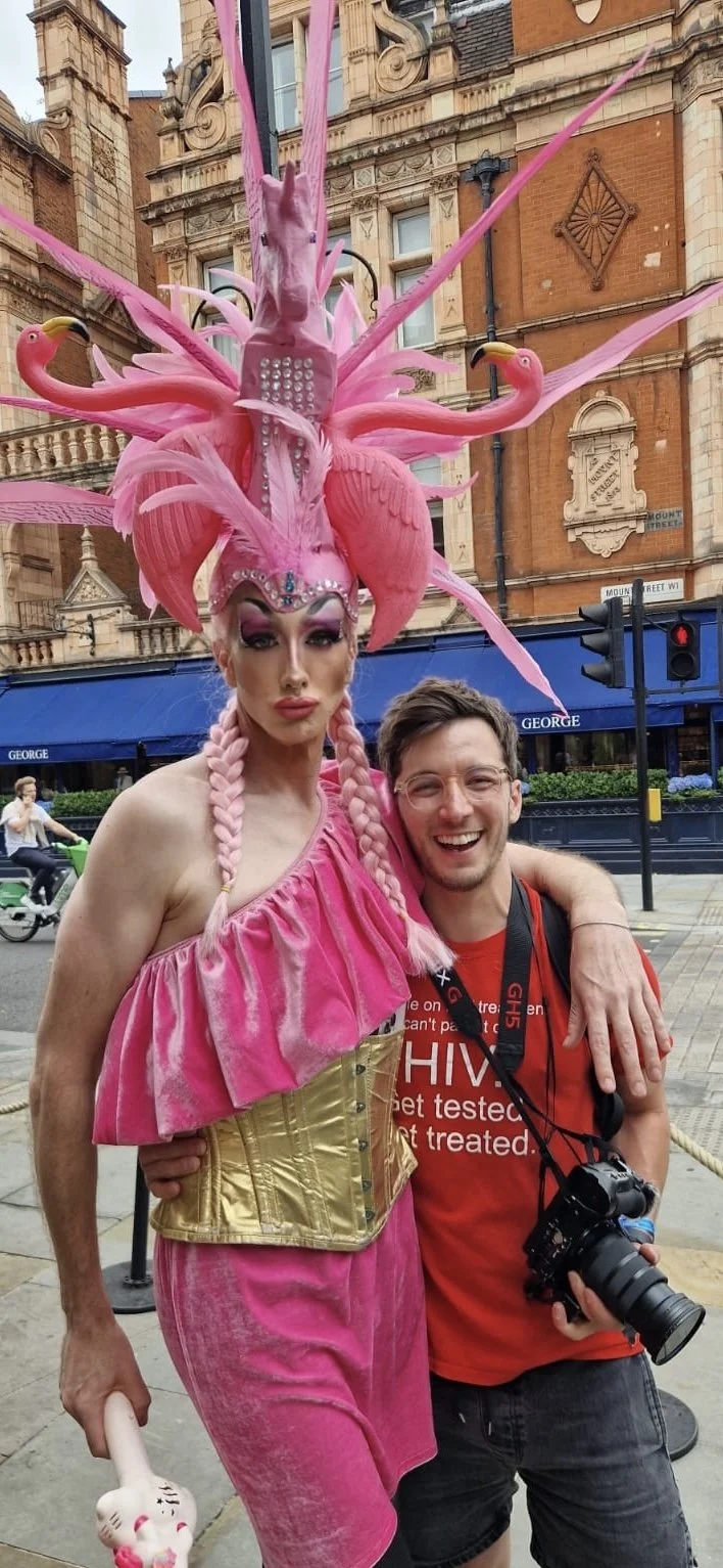 Drag performer in a pink outfit and an elaborate pink flamingo headdress with multiple heads and feathers posing with a smiling man in a red shirt, on a city street.
