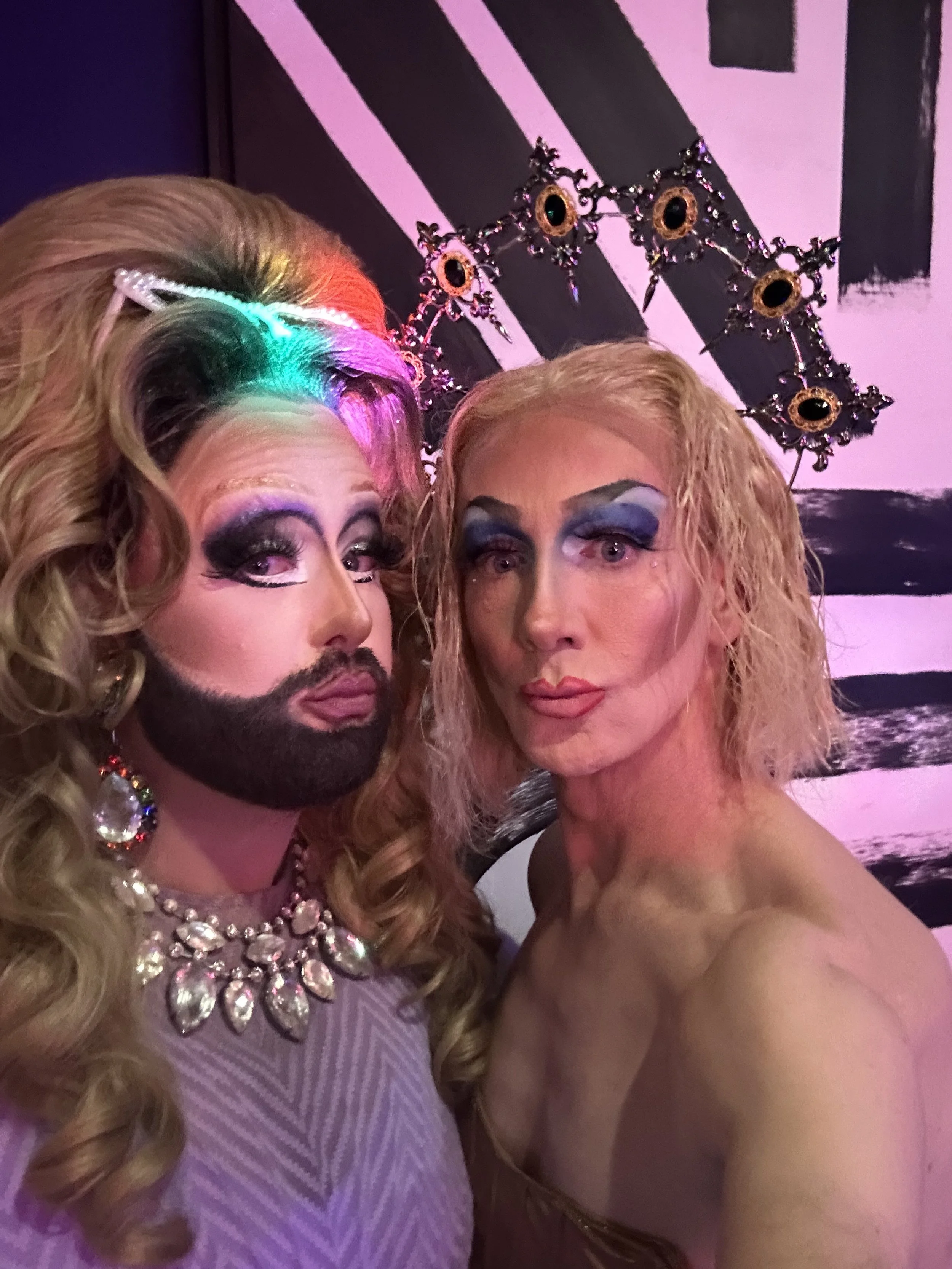 Two individuals with elaborate makeup and accessories posing together in a colorful setting. The person on the left has long curly blond hair, dramatic eye makeup, a bejeweled necklace, and a crown-like headpiece with large earrings. The person on th