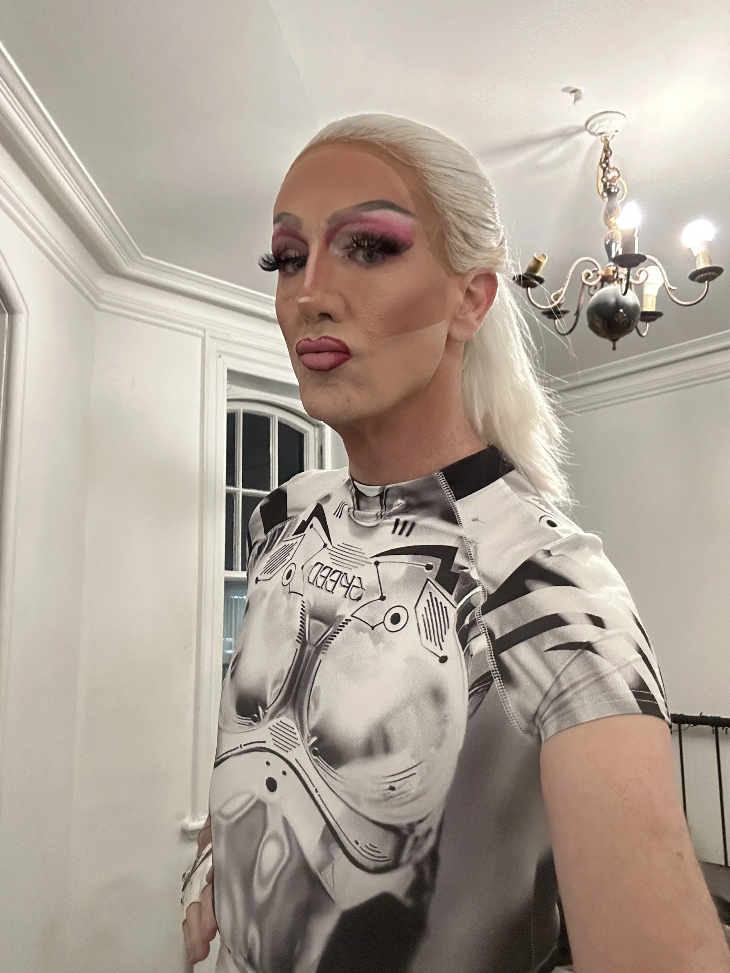 A person with platinum blonde hair, makeup featuring pink and purple eyeshadow, and dark eyeliner. They are wearing a fitted, short-sleeved, metallic gray shirt with a graphic print of a Stormtrooper helmet from Star Wars. The person has a confident 