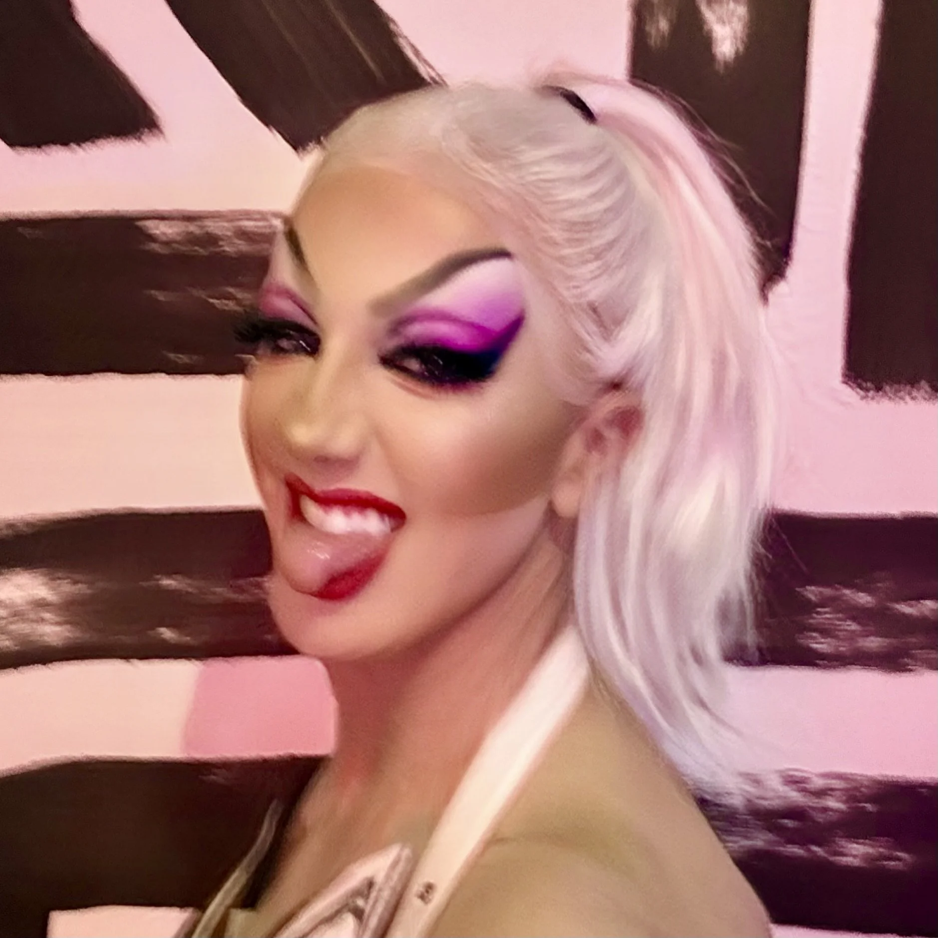 A woman with platinum blonde hair styled in loose waves, wearing vivid pink and purple eye makeup, red lipstick, sticking her tongue out, in front of a pink background with dark brown brush strokes.