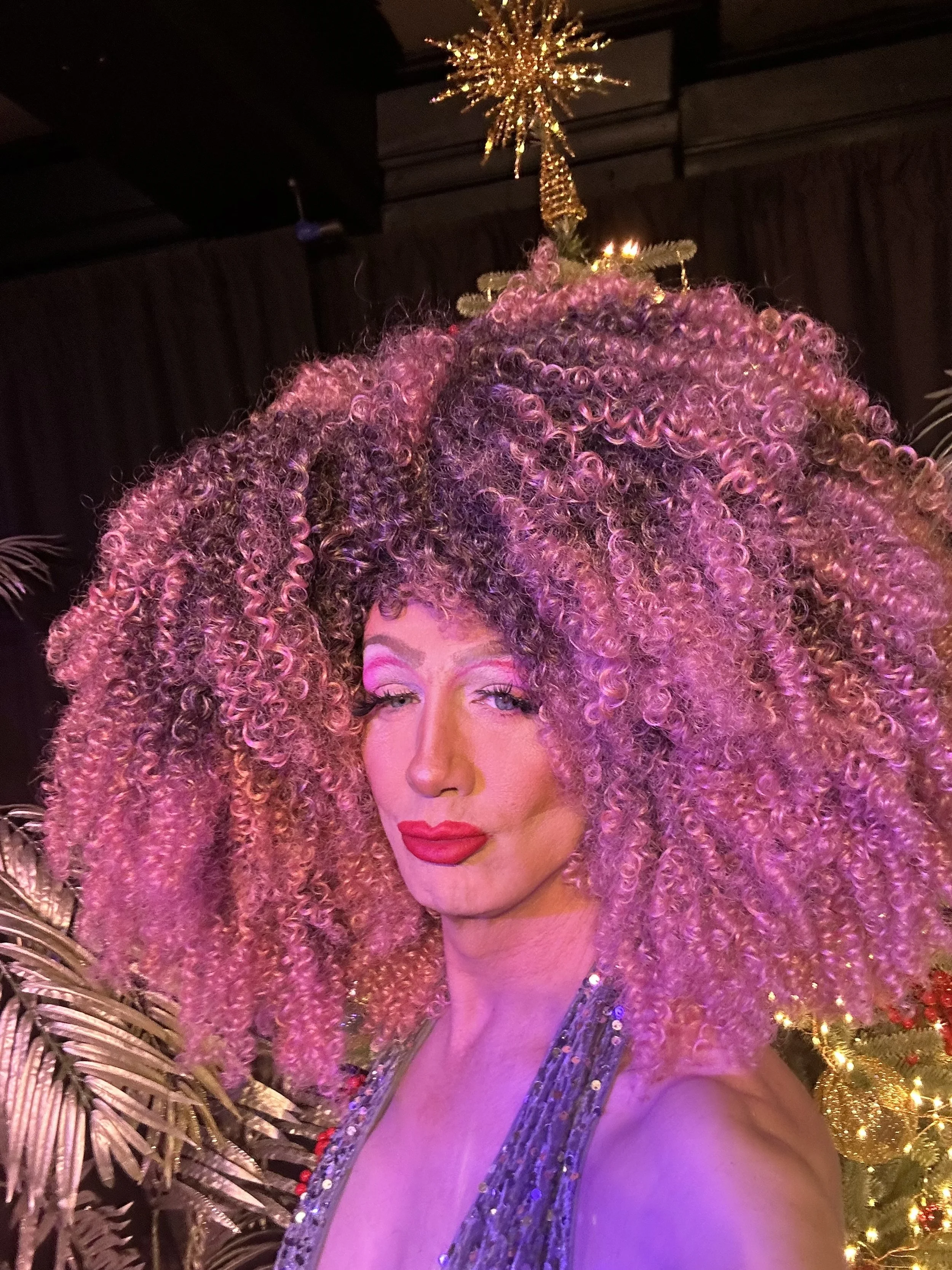 Person with voluminous, curly pink hair and bright makeup, standing in front of a Christmas tree with gold ornaments and lights.