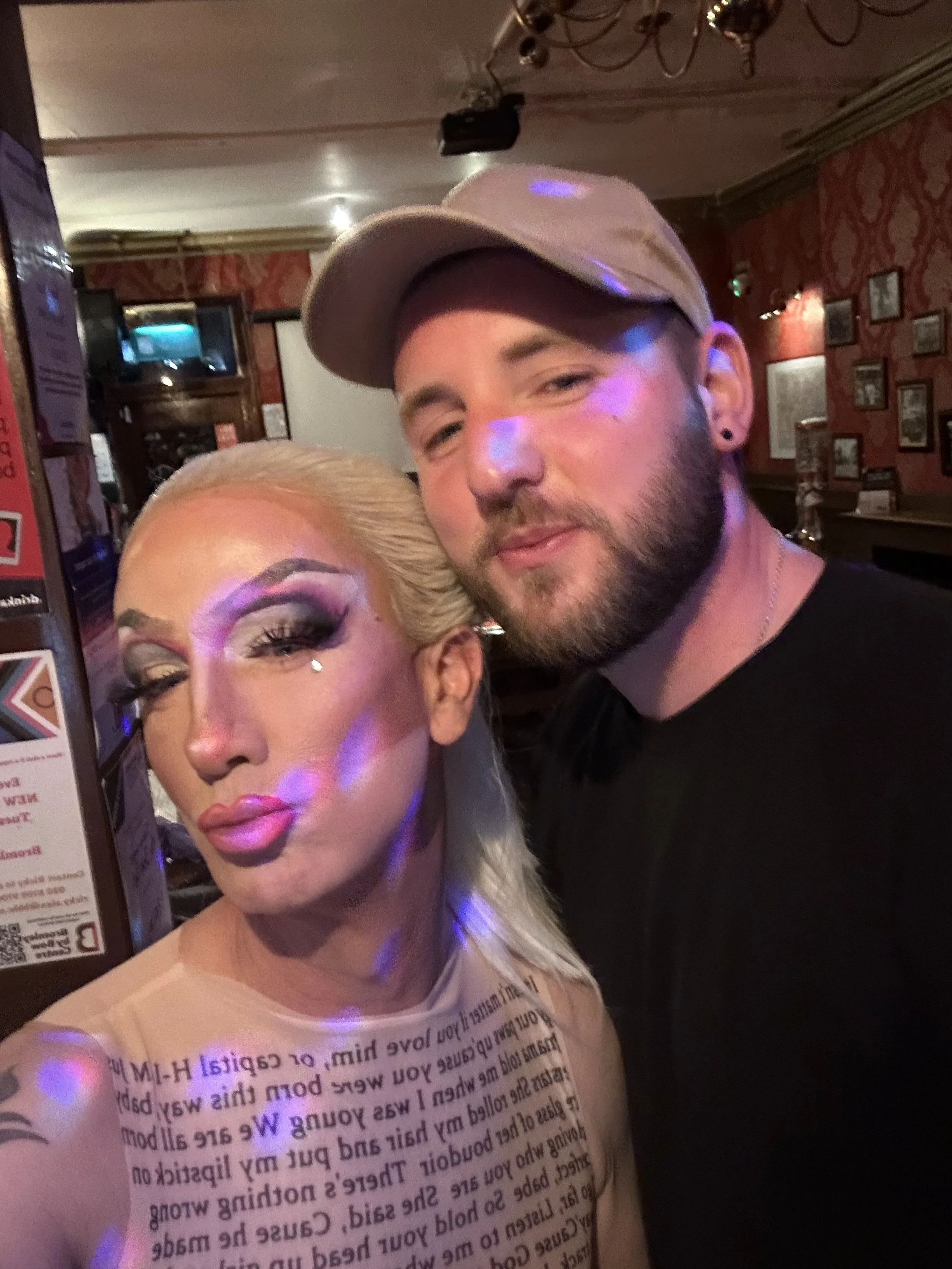 A woman with platinum blonde hair, intricate makeup, and a rhinestone on her cheek, taking a selfie with a man wearing a beige cap and black shirt inside a dimly lit bar or pub.