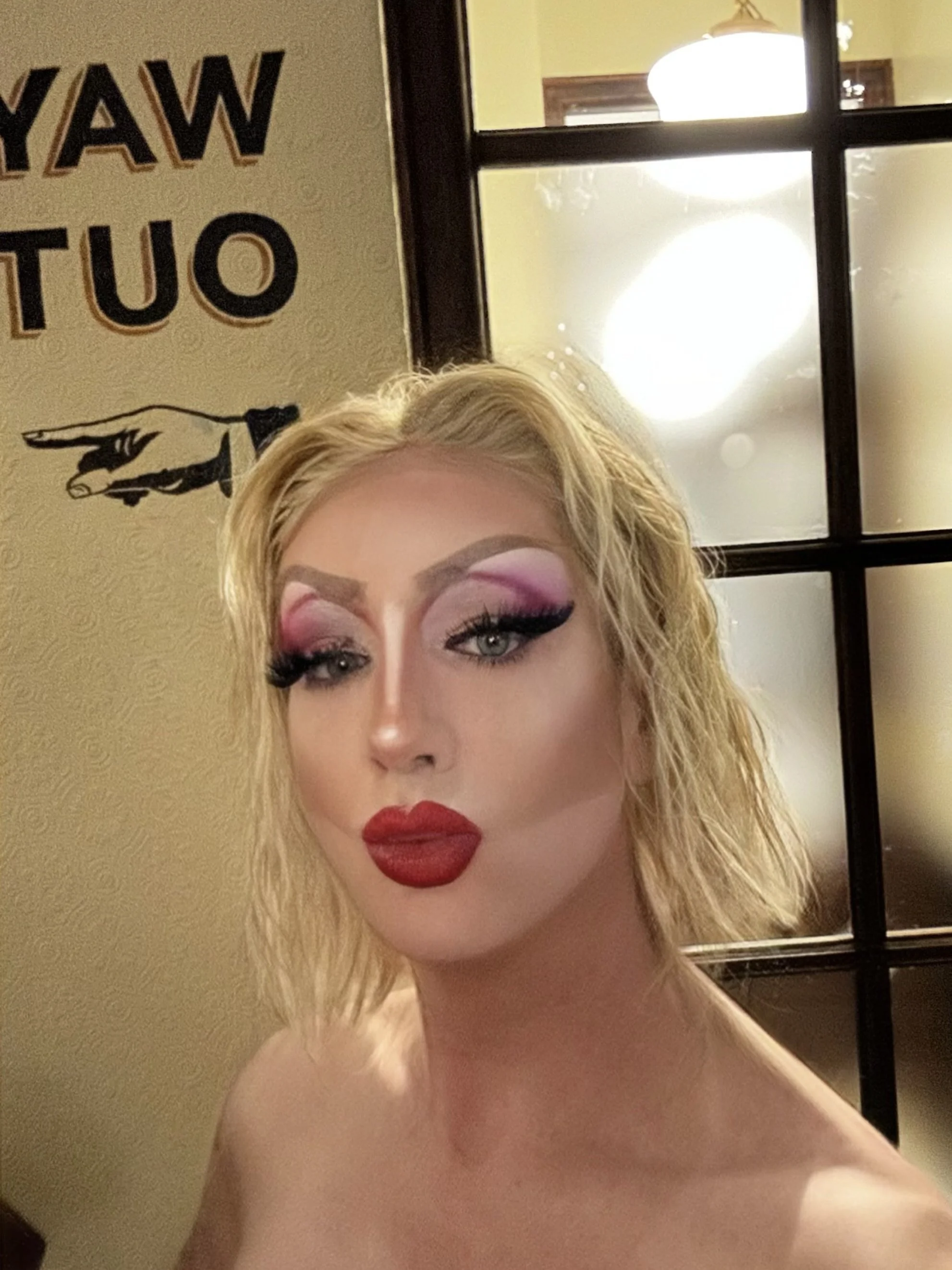 A person with blonde, curly hair, dramatic makeup including bold red lipstick, pink eyeshadow, and thick eyeliner, taking a selfie in front of a mirror.