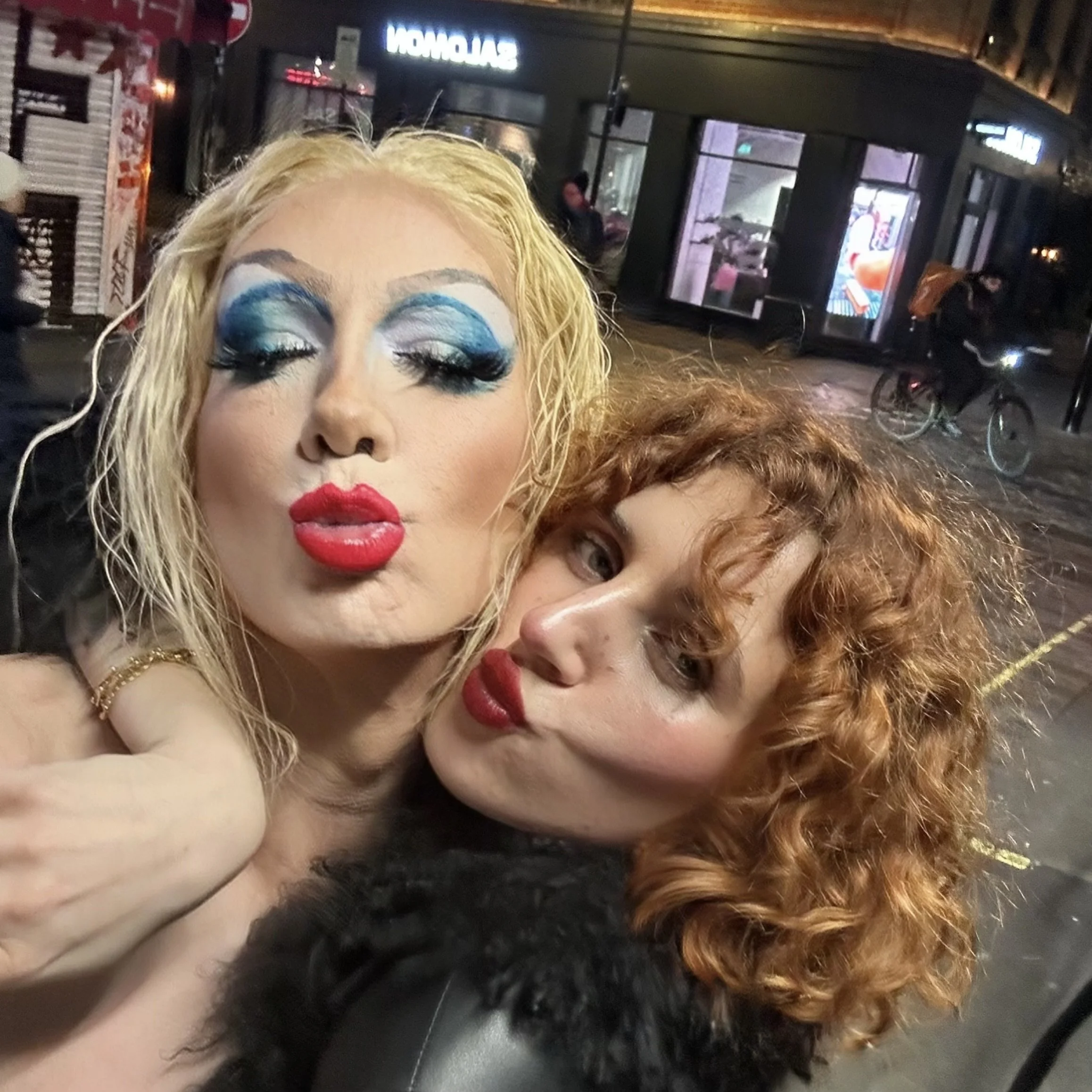 Two women taking a selfie outdoors at night. The woman on the left has blond curly hair, bold makeup with blue eyeshadow and red lipstick, and is making a kiss face. The woman on the right has red curly hair, lighter makeup with lipstick, and is lean
