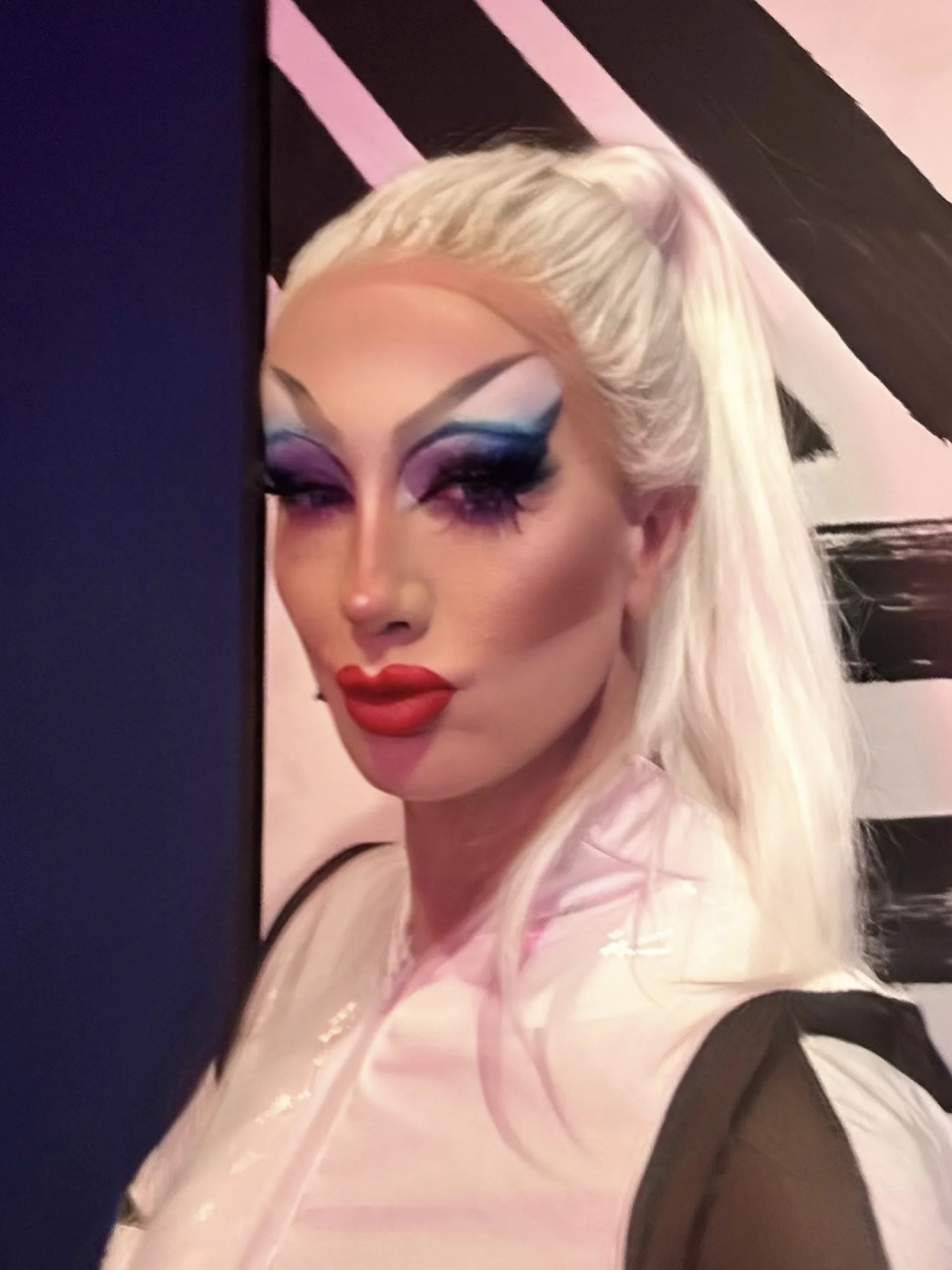 A person with platinum blonde hair styled in a high ponytail, wearing bold makeup with blue and purple eyeshadow, thick eyeliner, dramatic lashes, and bright red lipstick, posing against a black and pink striped background.