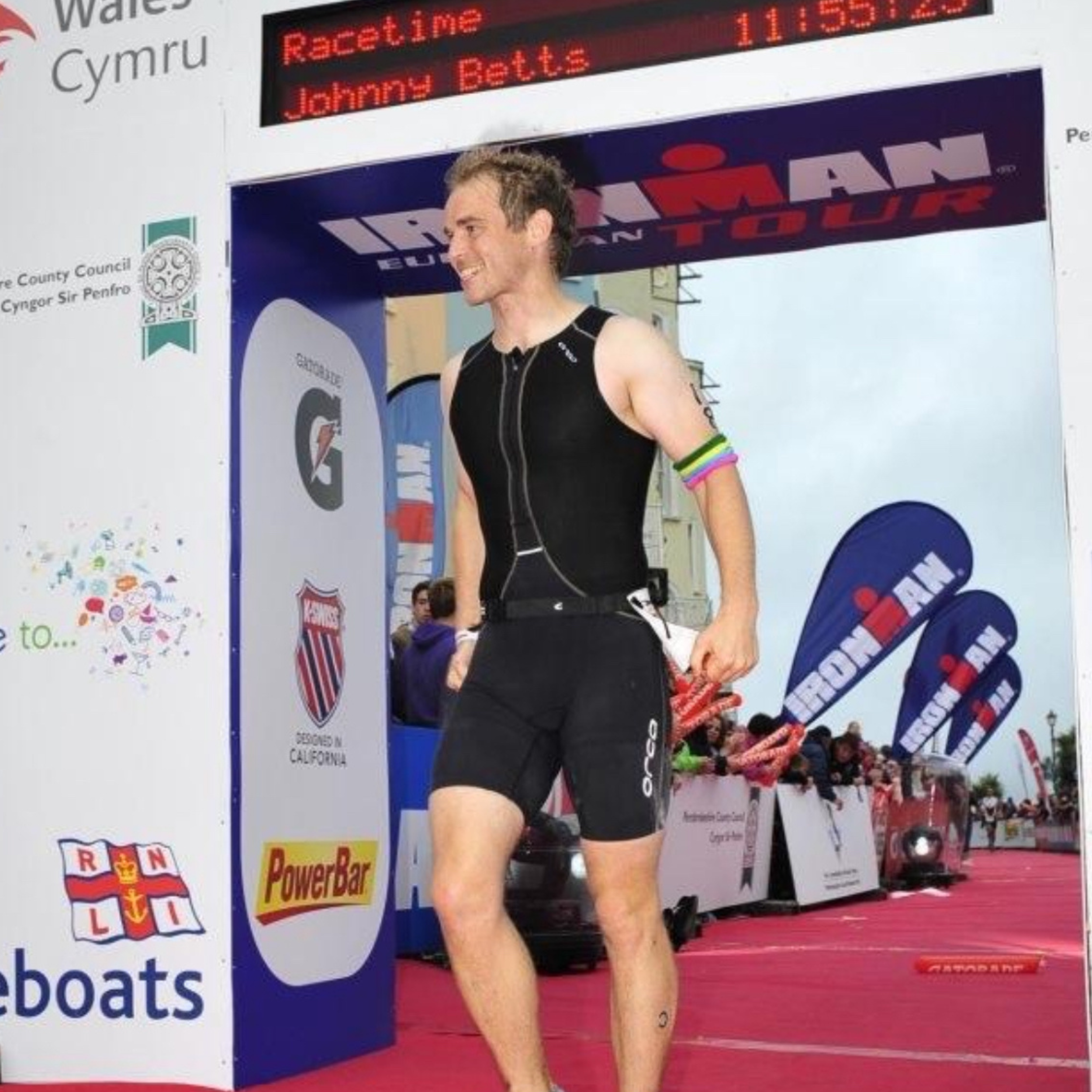 A male athlete in a black triathlon suit standing at the finish line of an Ironman event, with race banners and flags in the background.