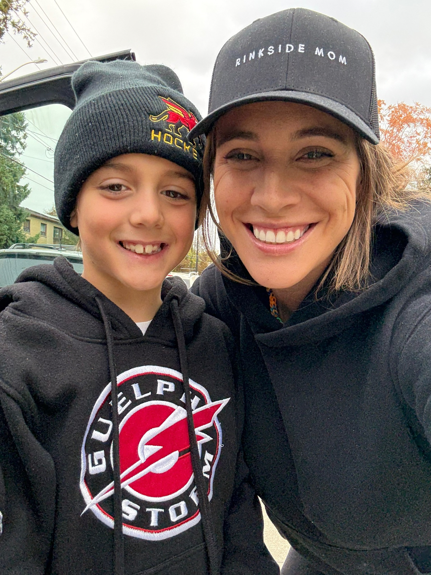 A woman and a young boy smiling and taking a selfie outdoors. The woman is wearing a black cap with 'RINKSIDE MOM' written on it, and the boy is wearing a black beanie and a black hoodie with a logo that says 'GUILDER STO' and features a red lightning bolt and a hockey puck.