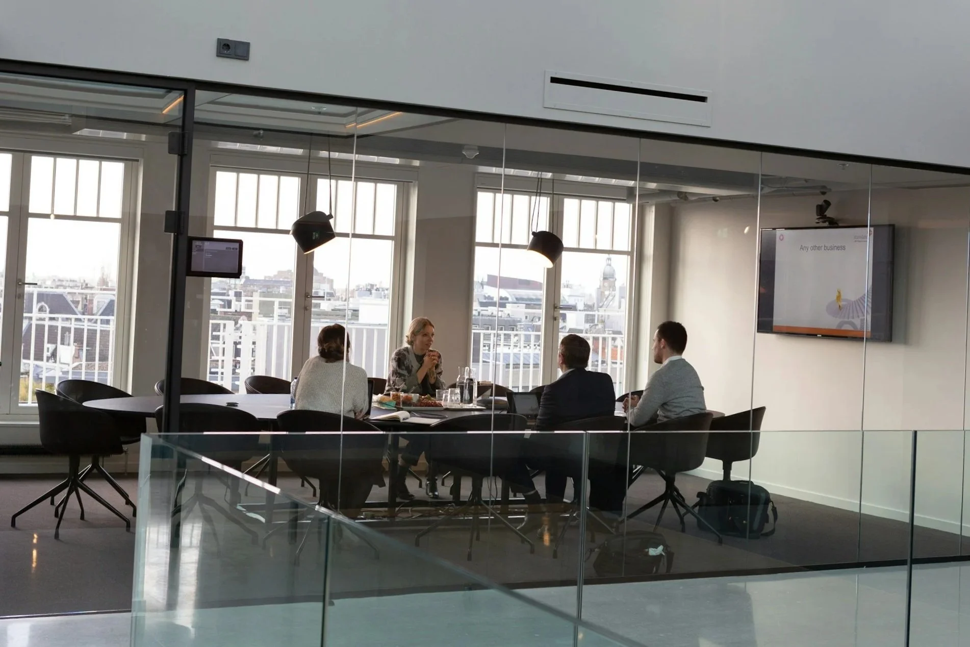 Four people seated around a conference table in a modern office meeting room with large windows and city skyline view, engaged in discussion.