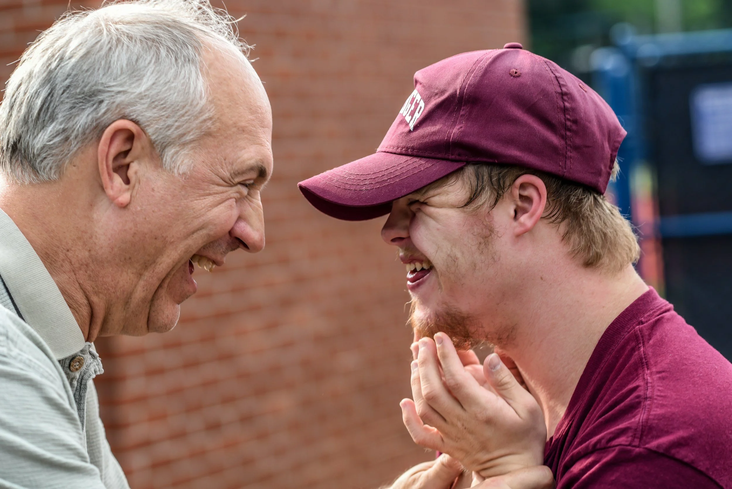 Two men laughing close together outside, one older with gray hair and the other younger with a beard and wearing a maroon cap.