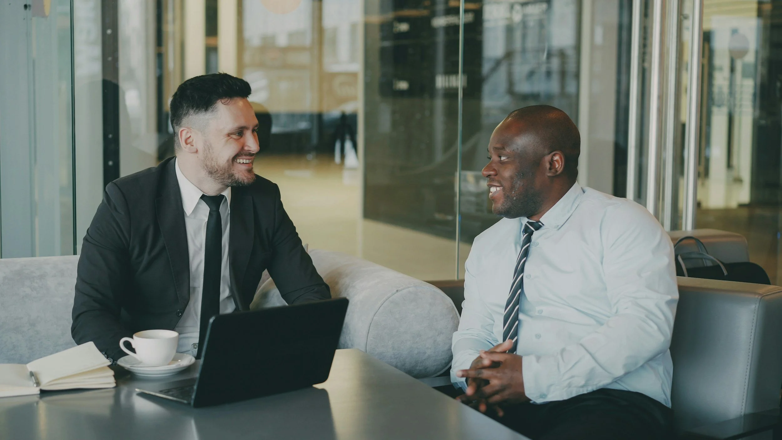 Two businessmen having a friendly conversation in a modern office lounge, with a laptop, notebook, and coffee cup on the table.