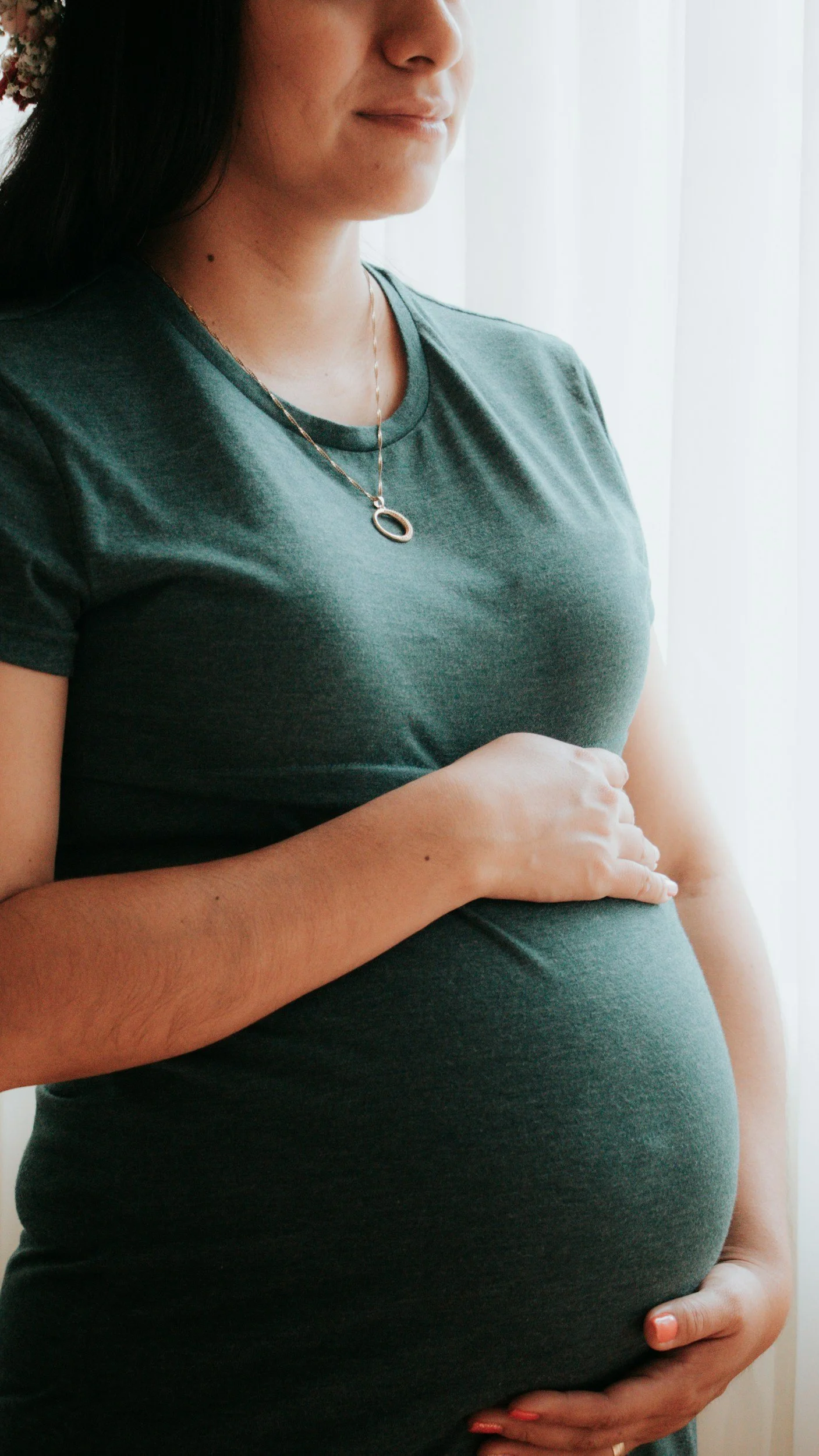 A pregnant woman in a green T-shirt gently holding her belly, standing near a window with white curtains, wearing a necklace with a circular pendant.