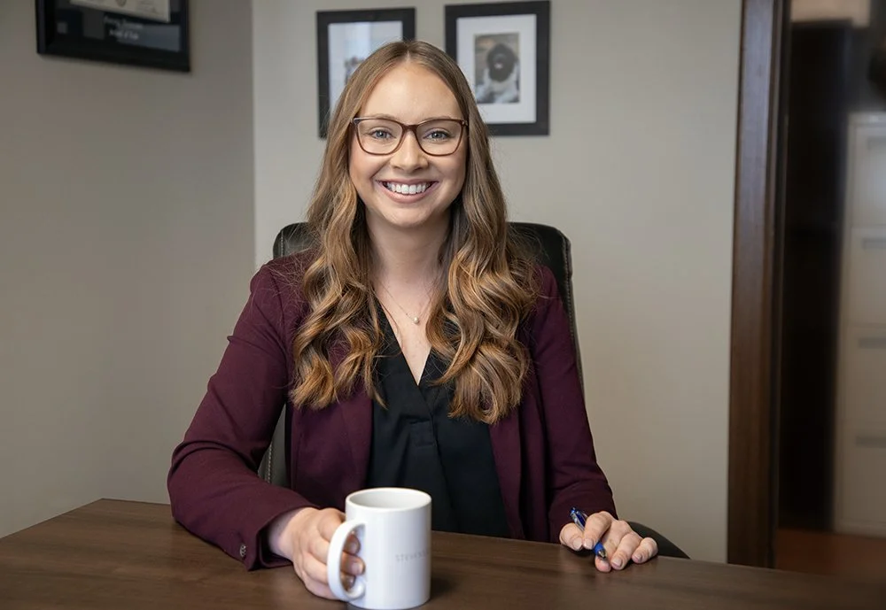 A smiling woman with long wavy hair, wearing glasses and a burgundy blazer, sits at a wooden desk holding a white coffee mug.