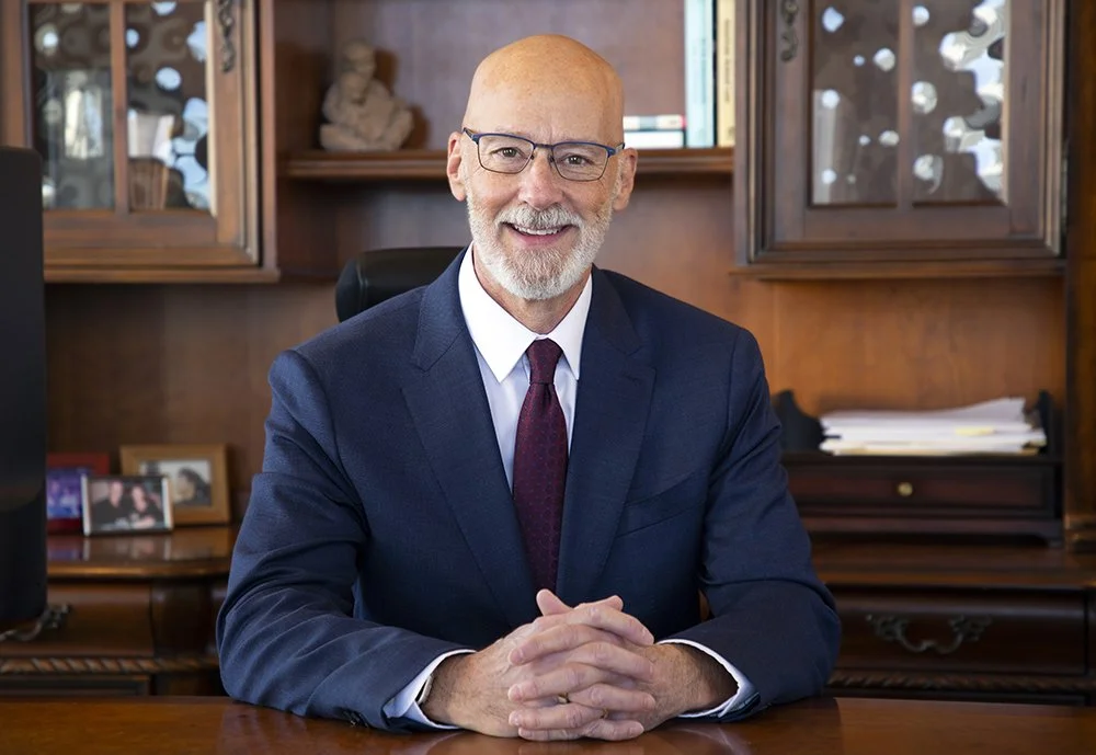 A middle-aged man with a beard and glasses, dressed in a navy suit, white shirt, and maroon tie, sitting at a wooden desk in an office, smiling at the camera.