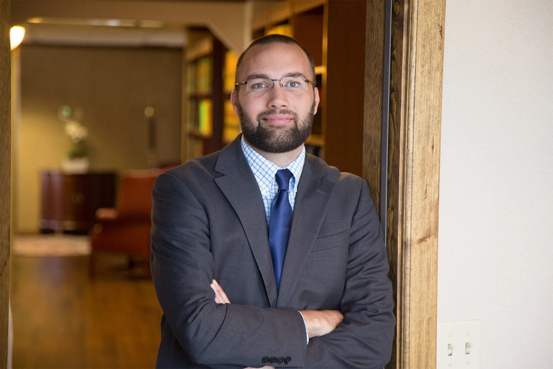 A man with a beard and glasses wearing a dark gray suit, blue checkered shirt, and blue tie standing with arms crossed inside a room with a wooden doorway, bookshelves, and furniture in the background.