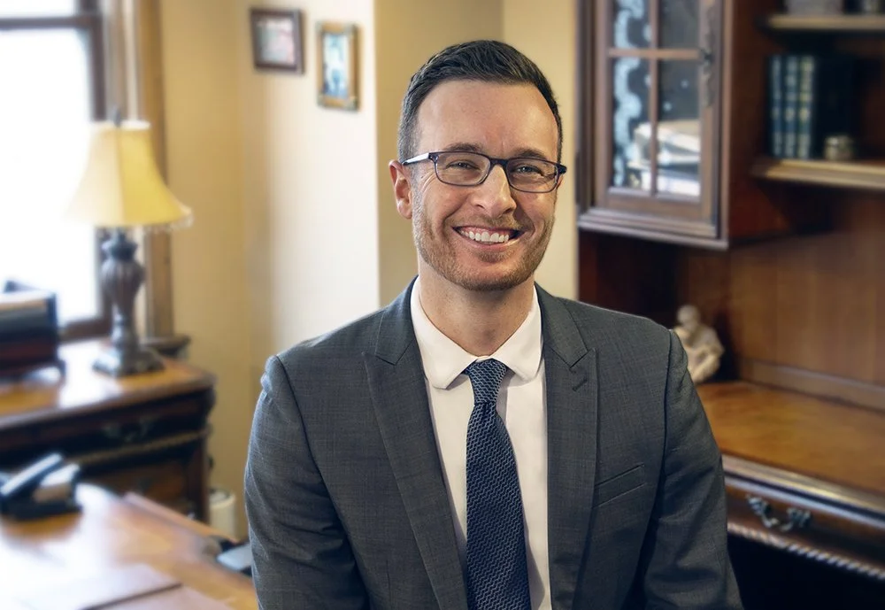 A smiling man in a gray suit, white shirt, and blue tie, wearing glasses, sitting in an office with wooden furniture, a window, books, and framed pictures in the background.