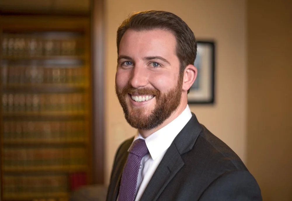 A smiling man in a suit with a beard and mustache, standing in a room with a bookshelf and framed picture in the background.