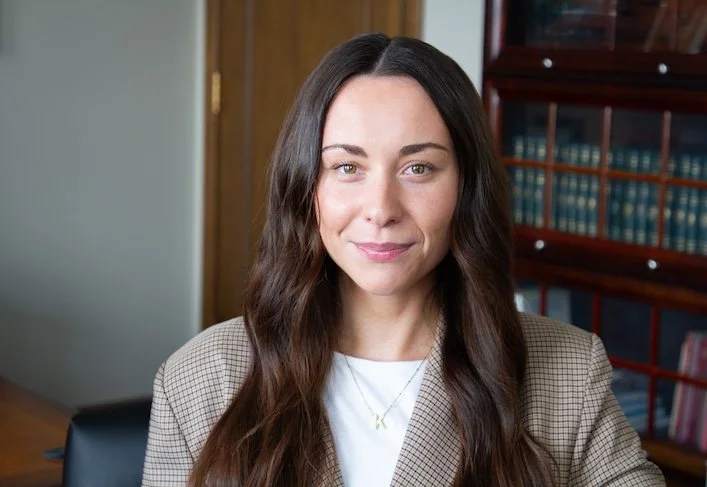 A woman with long brown hair and light skin, sitting in an office with wooden bookshelves and a closed wooden door in the background.