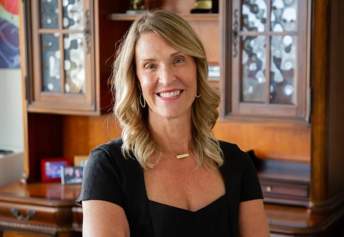 A smiling woman with shoulder-length blonde hair, wearing a black top with a square neckline, gold jewelry, and earrings, standing in a room with a wooden cabinet behind her.