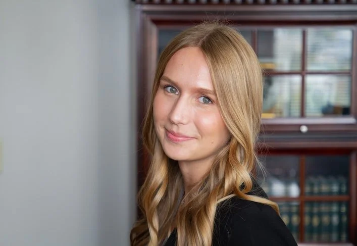 A young woman with long blonde hair and blue eyes smiling softly, standing in front of a bookcase in a room with a window.