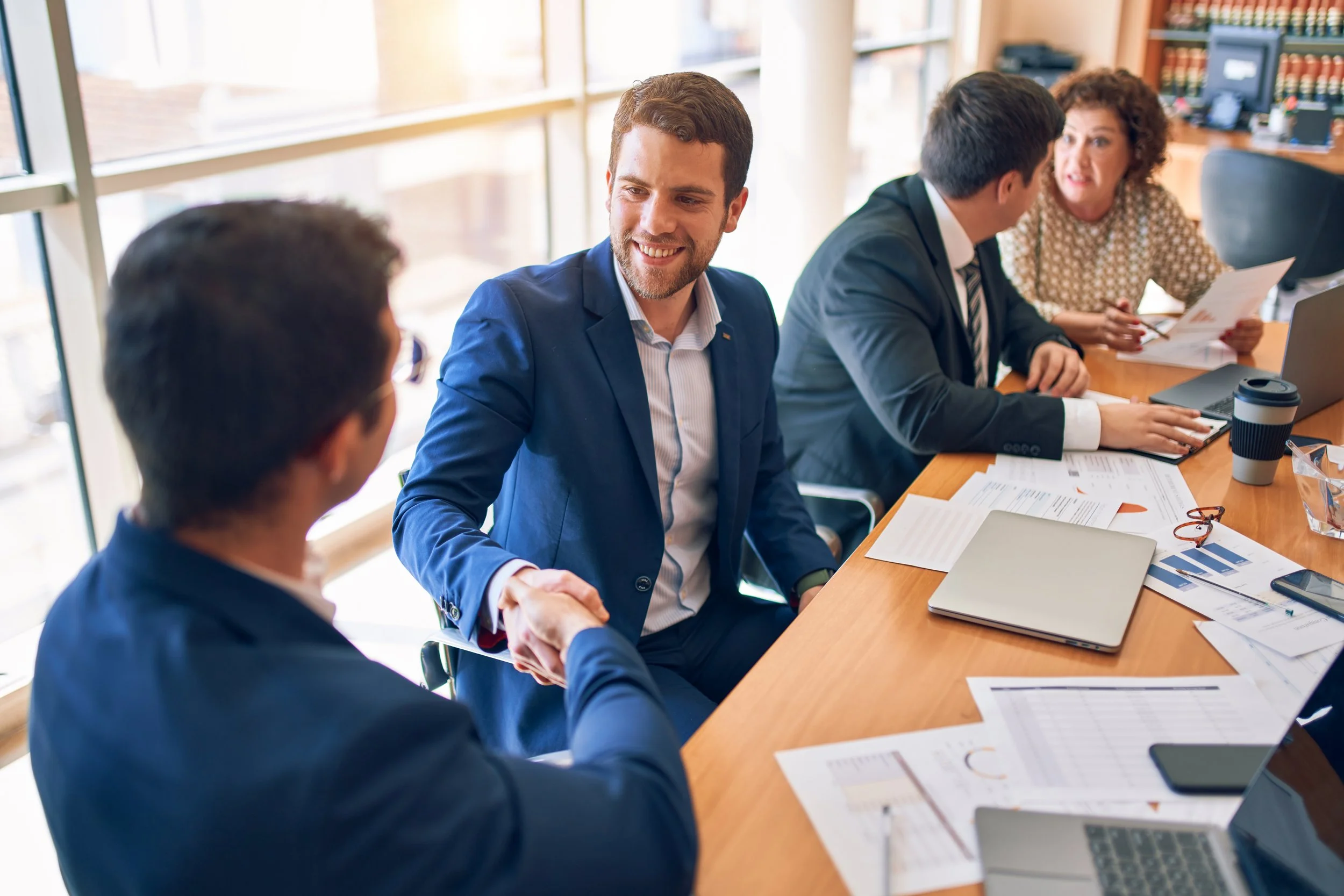 Business professionals in suits sitting around a table in a bright office, shaking hands and discussing documents.