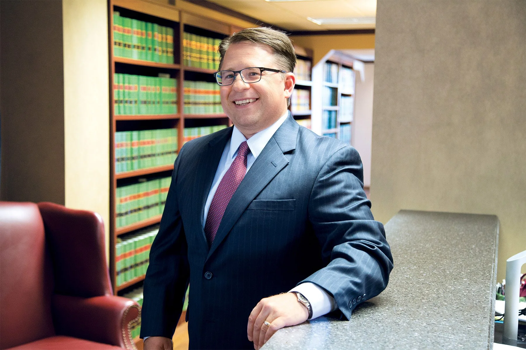 A man in a business suit smiling, standing in a library or office with shelves of green books behind him.
