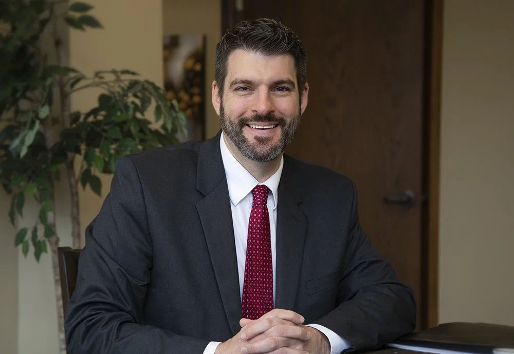 A man in a dark suit and red tie sitting at a table in an office, smiling.
