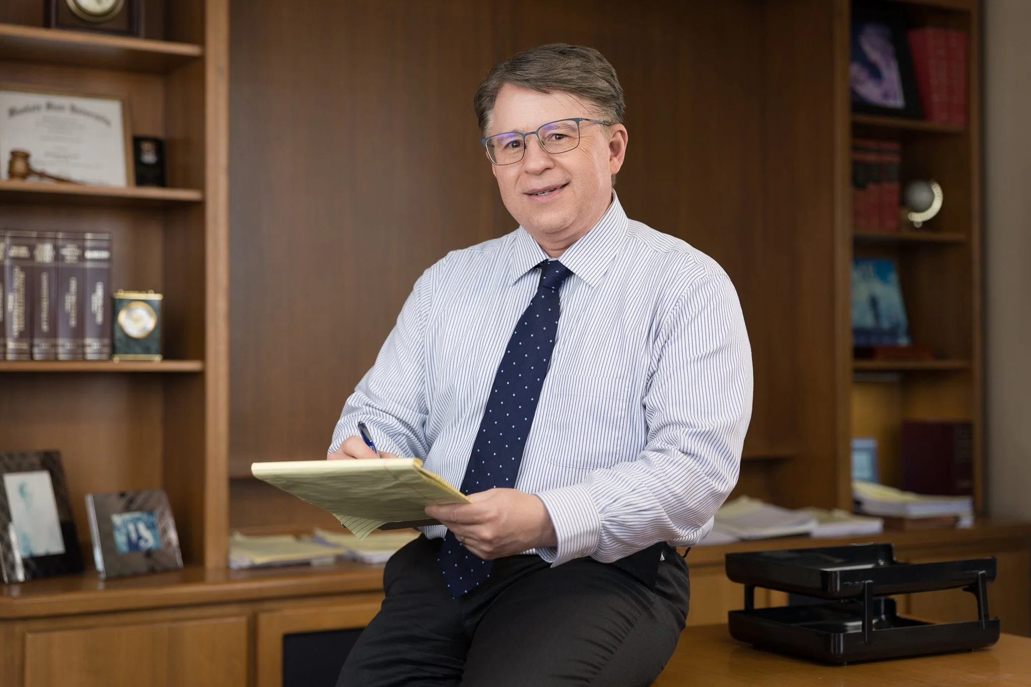Man in tie and glasses sitting on corner of desk with pad of paper in hand