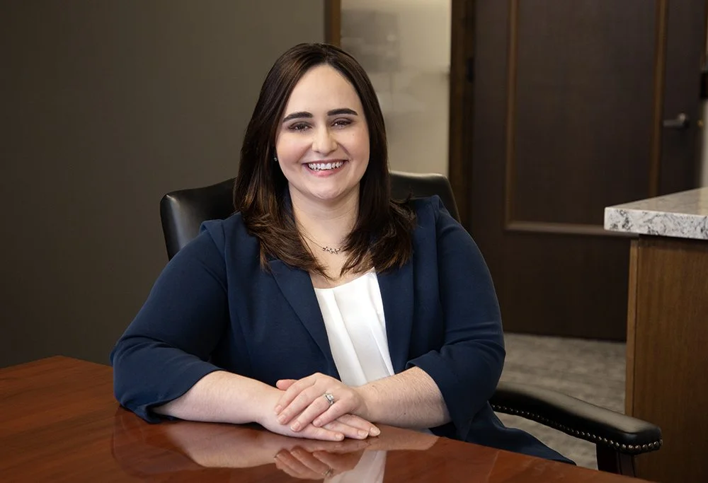 A woman with dark brown hair, smiling, sitting at a wooden table in an office, wearing a navy blazer and white blouse.