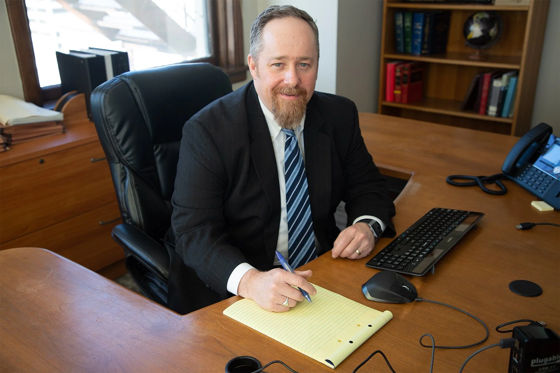 A man in a black suit and striped tie sitting at a wooden desk, holding a pen over a yellow notepad, with a computer keyboard, mouse, and office phone on the desk.