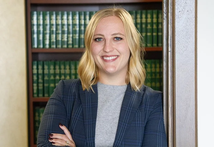 A smiling woman with blonde hair, wearing a gray shirt and plaid blazer, standing in front of a bookshelf filled with green and gold books.