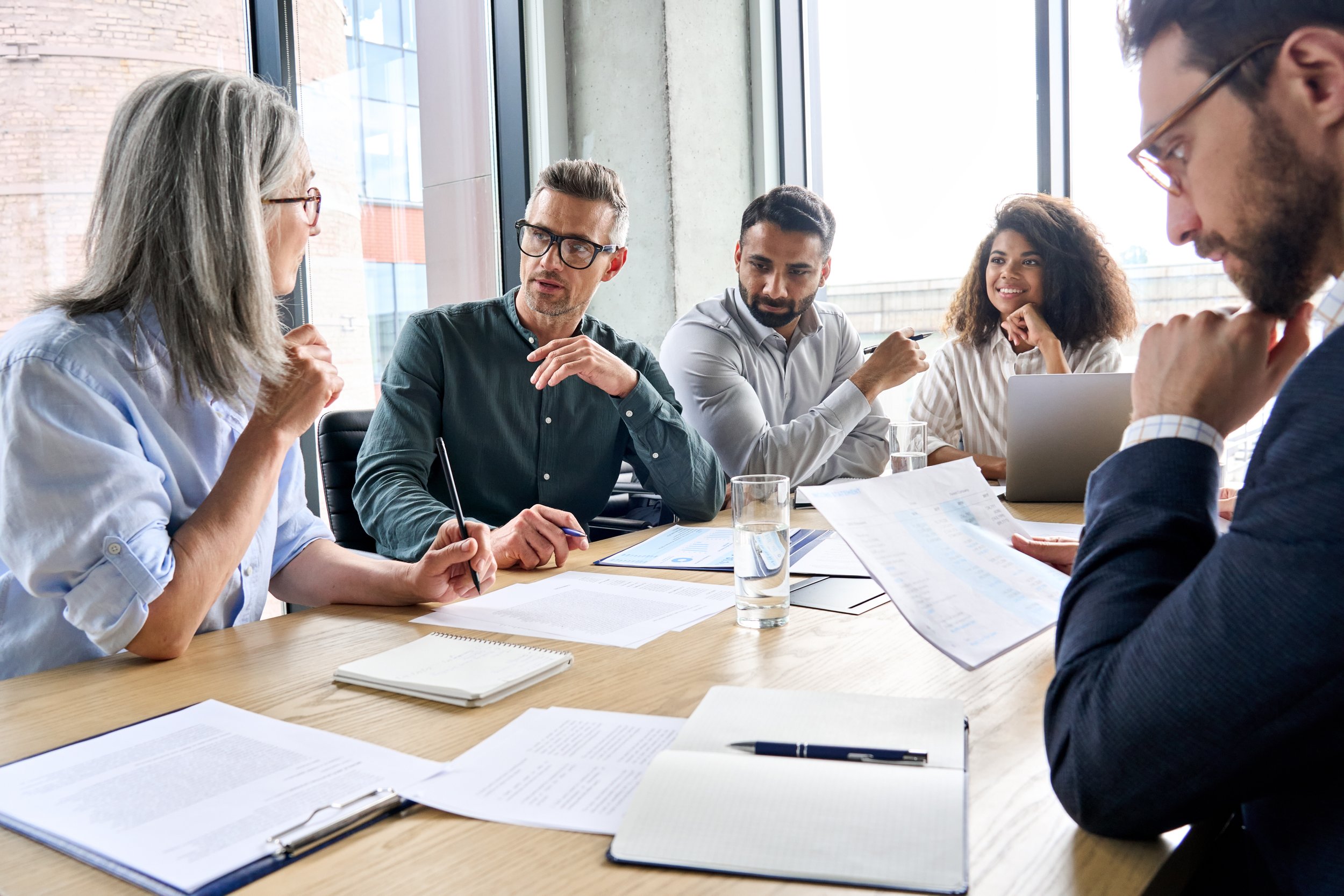 Group of five diverse professionals having a discussion in a modern conference room with large windows.