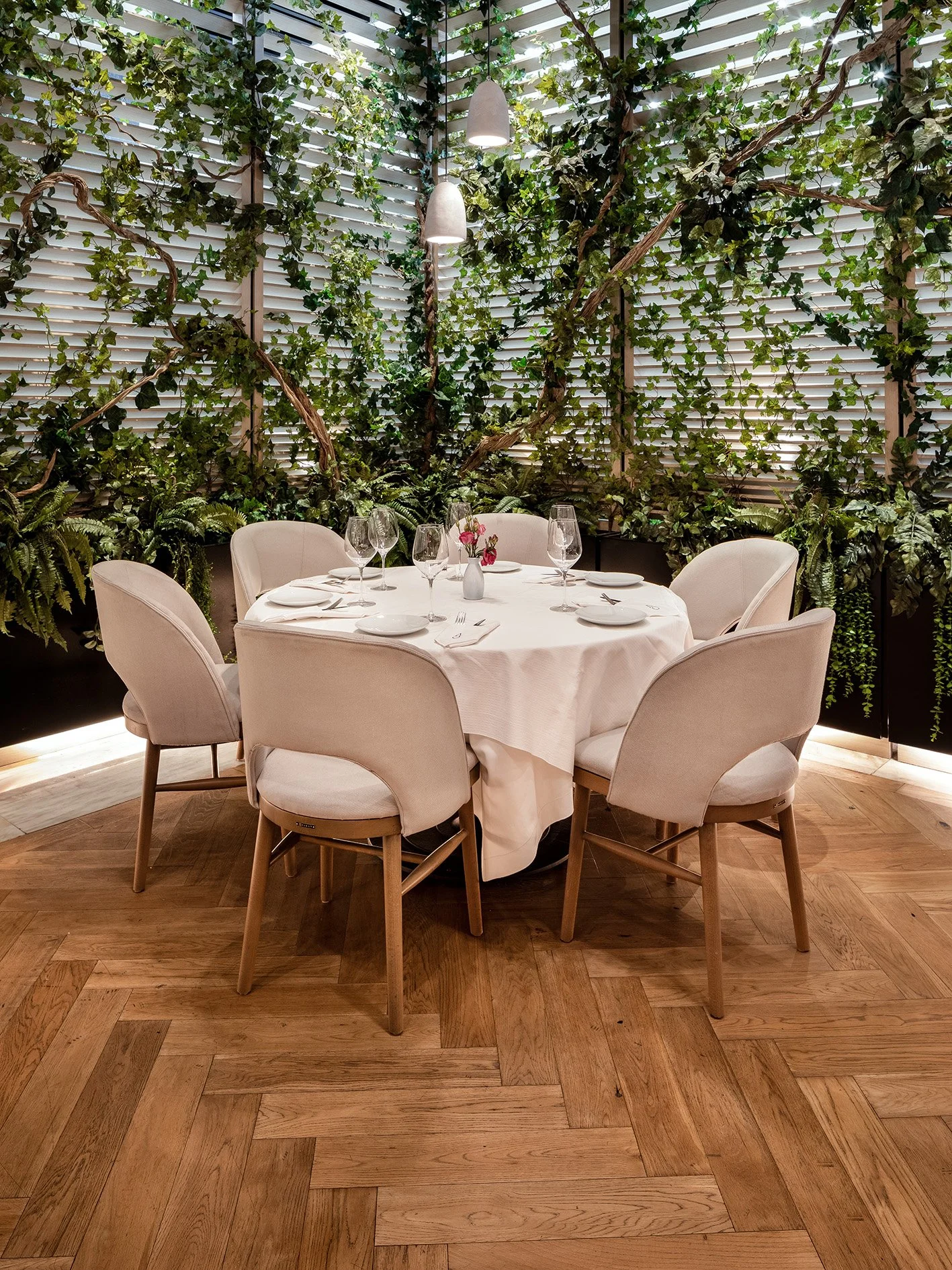 Mesa redonda con mantel blanco, seis copas y vajilla, en un restaurante con paredes cubiertas de plantas y ventanas con persianas blancas. 