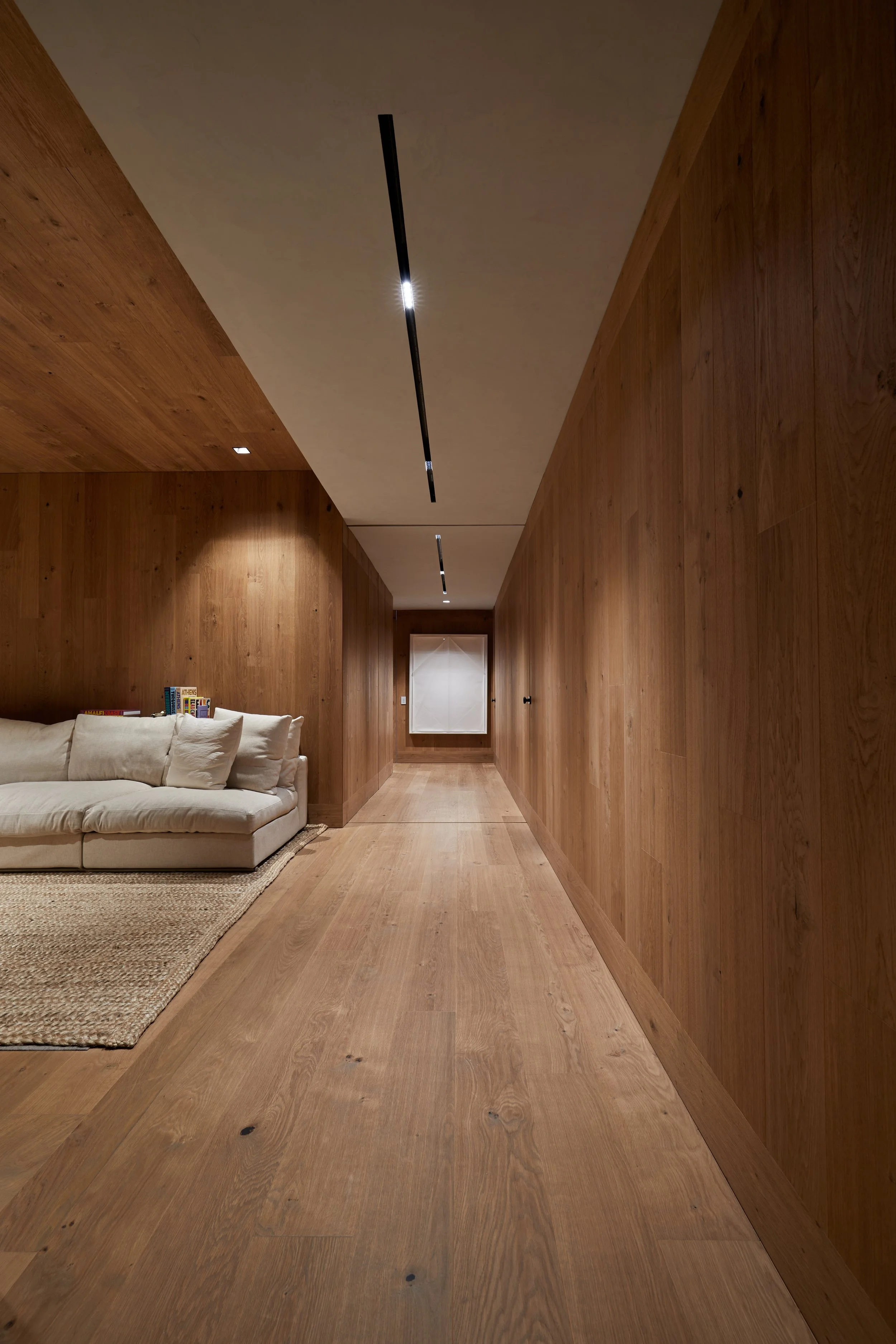Pasillo de techo alto con paredes y suelo de madera, iluminado por luces empotradas, con un sofá blanco y una alfombra en el lado izquierdo, y una ventana en la pared al fondo.