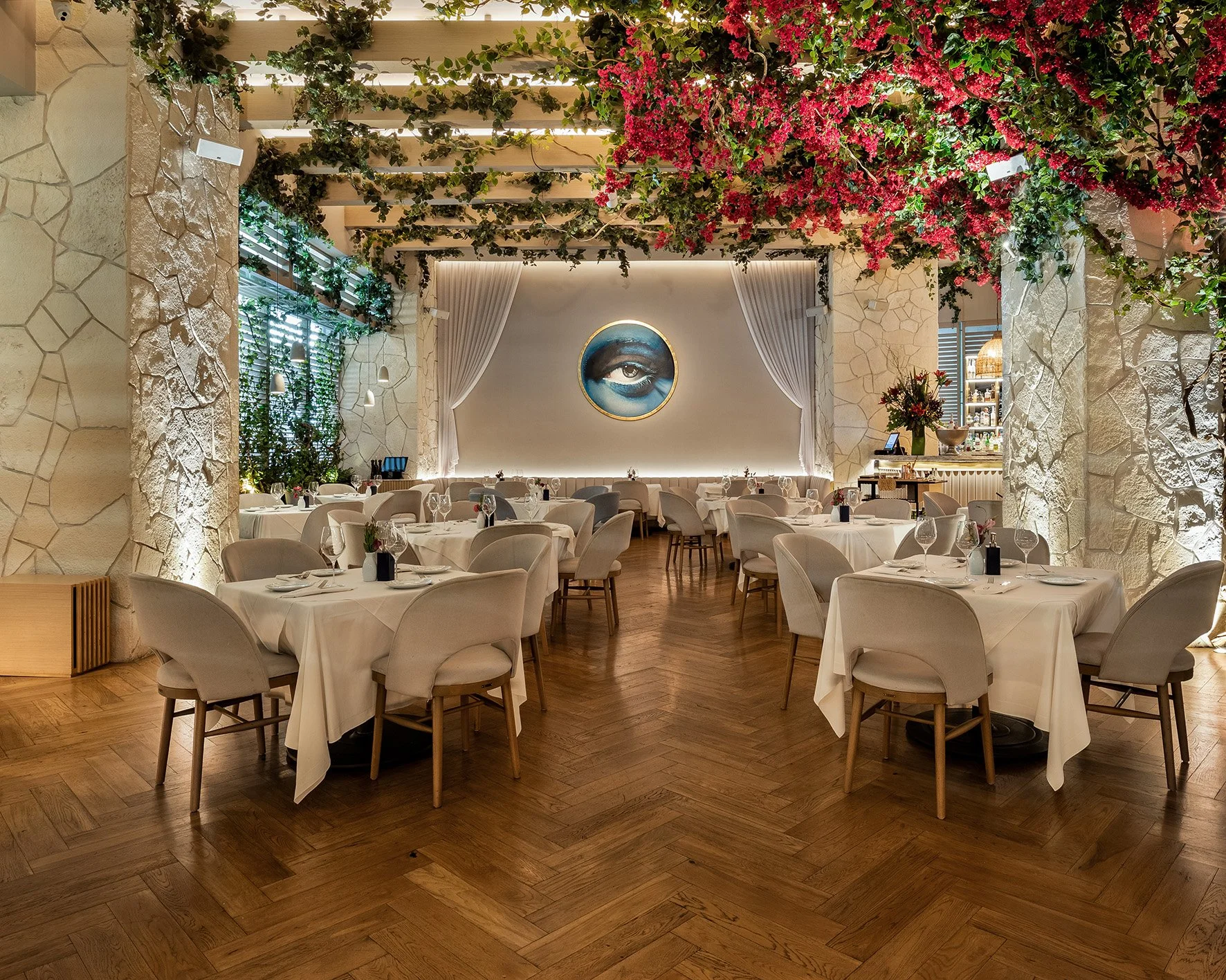 Restaurante elegant con mesas cubiertas con manteles blancos, decorado con plantas y flores, con un mural de un ojo en el fondo y un techo adornado con enredaderas y flores color rosa.