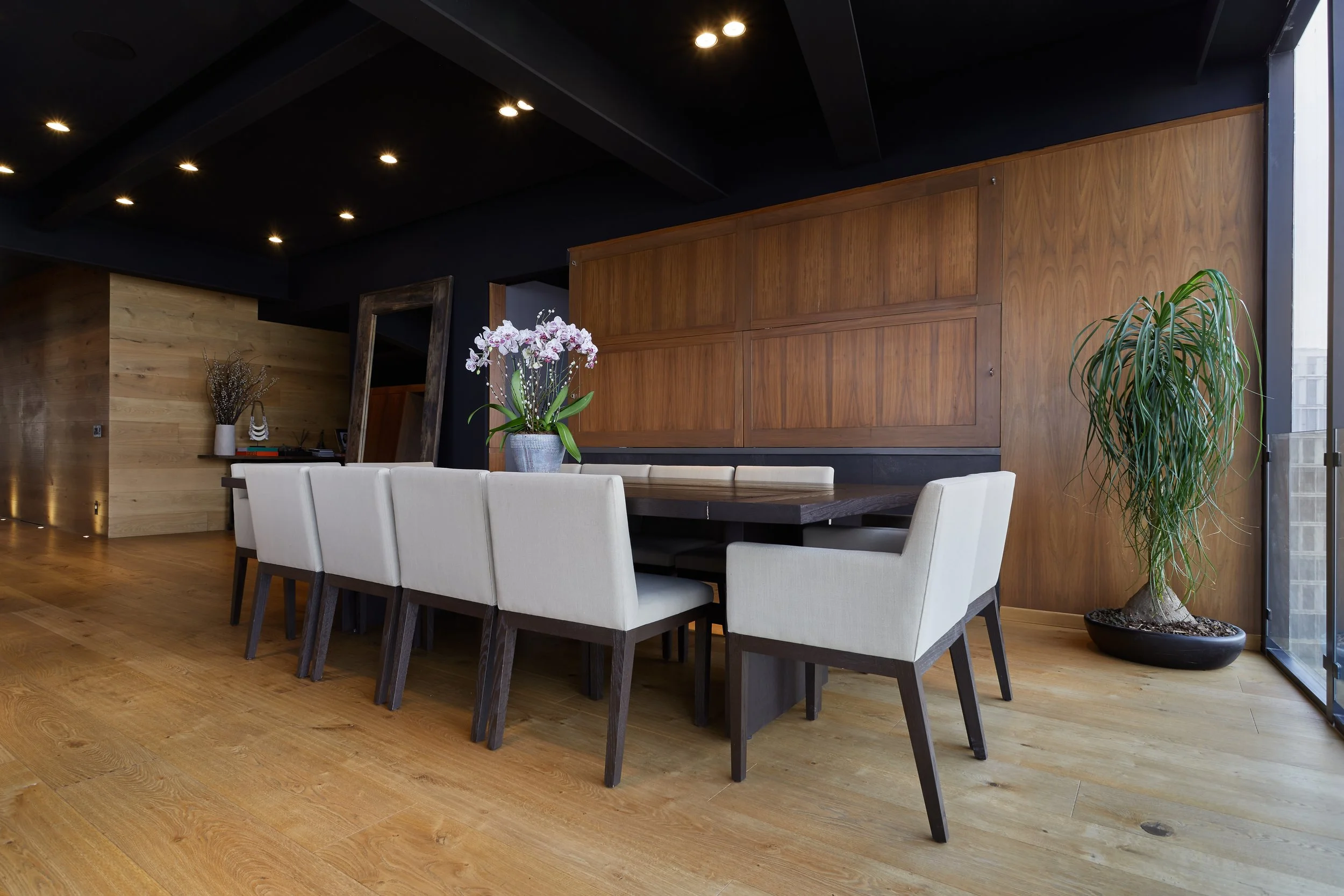 Una moderna sala de comedor con una mesa de madera y sillas tapizadas en blanco. Hay una planta grande en un recipiente negro y un florero con flores en la mesa. La pared de fondo está cubierta de madera y el techo es negro con luces empotradas.