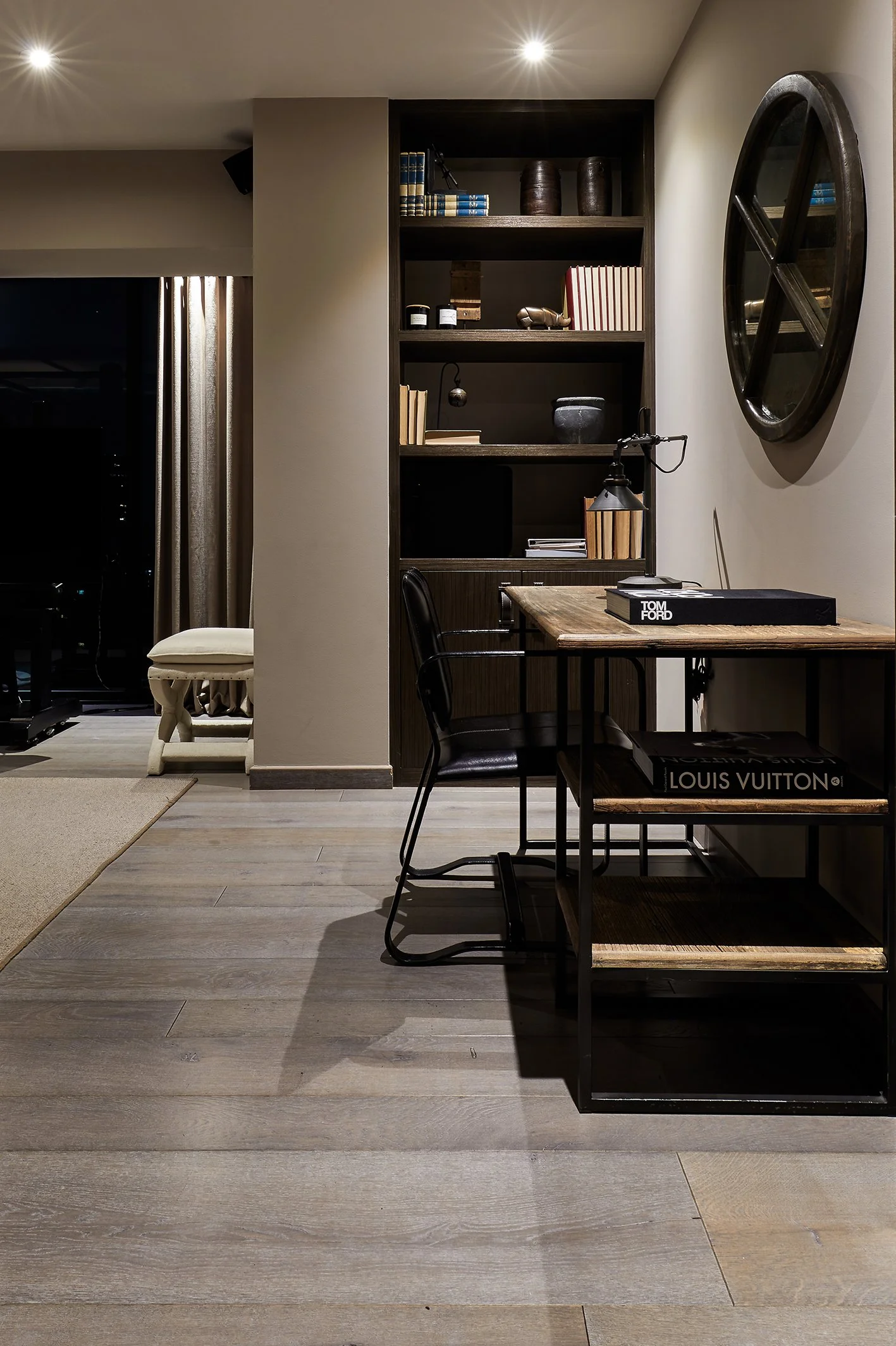 Estudio en una habitación con estantería de madera con libros y objetos decorativos, escritorio de madera, silla negra y pared con espejo redondo.