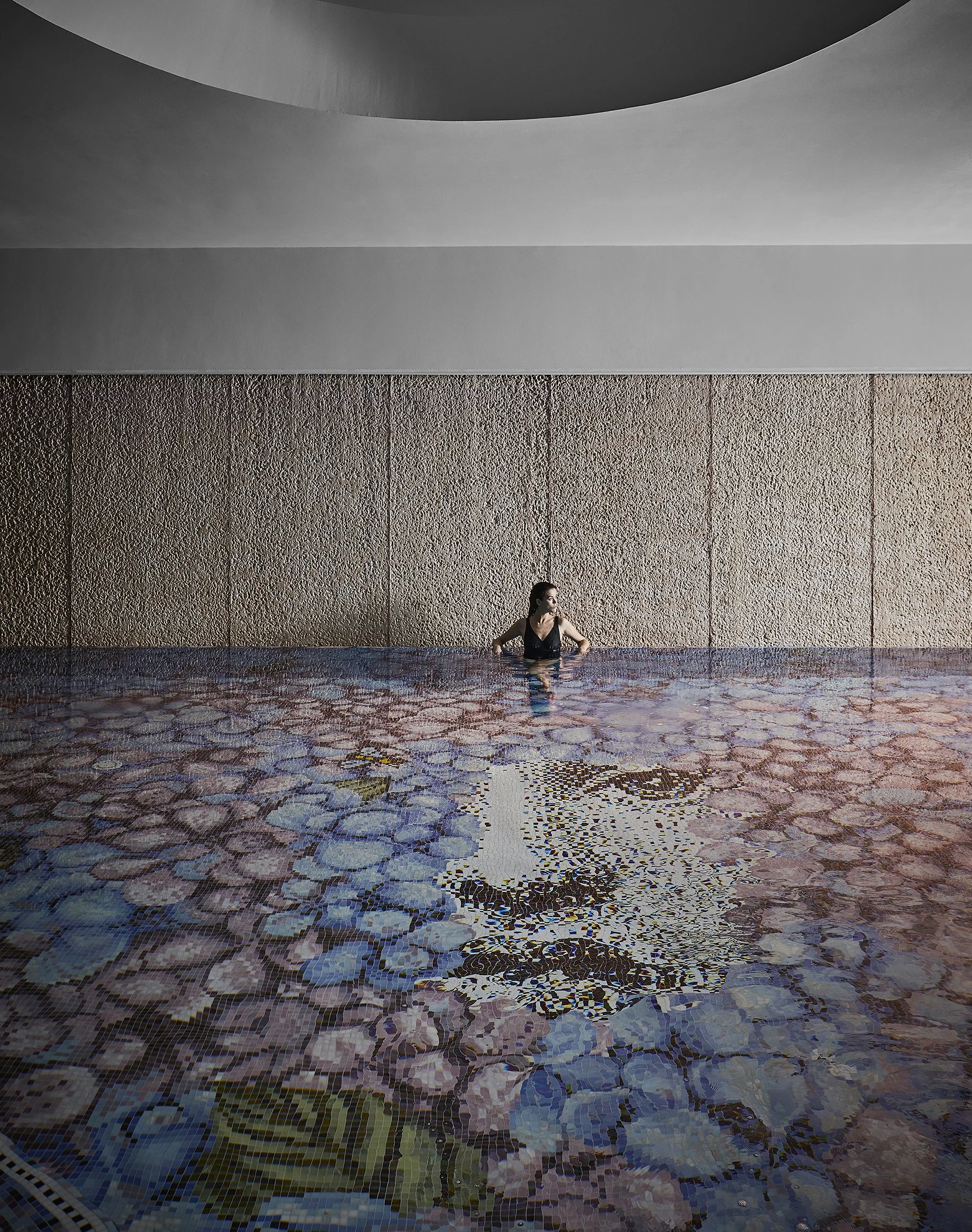 Una mujer en una piscina interior con paredes de textura rugosa y un techo curvado, la piscina tiene un mosaico con diferentes colores y un diseño de flor o figura abstracta.