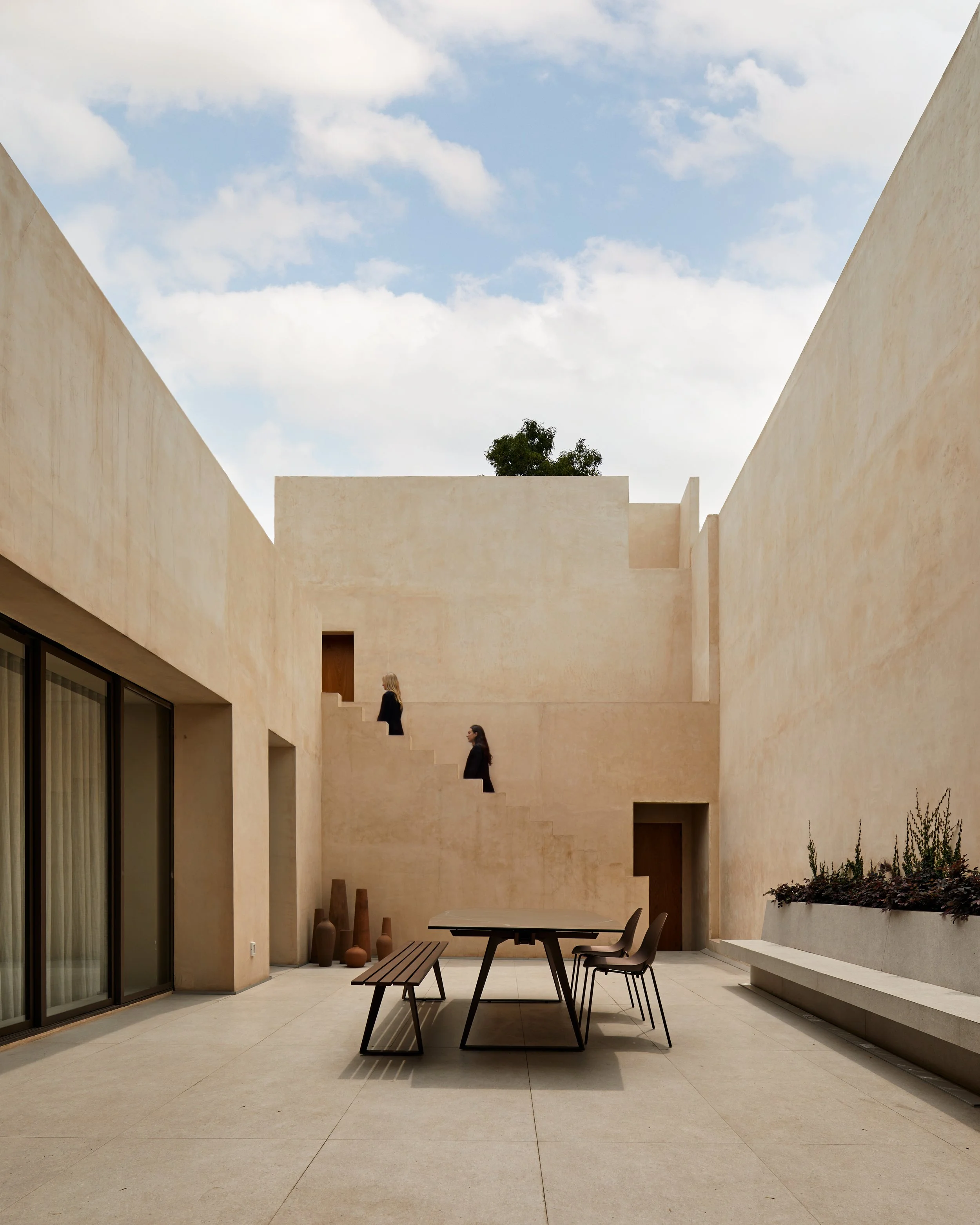 Patio moderno con paredes de tono beige, mobiliario de madera y metal, plantas en macetas y dos personas caminando por una escalera