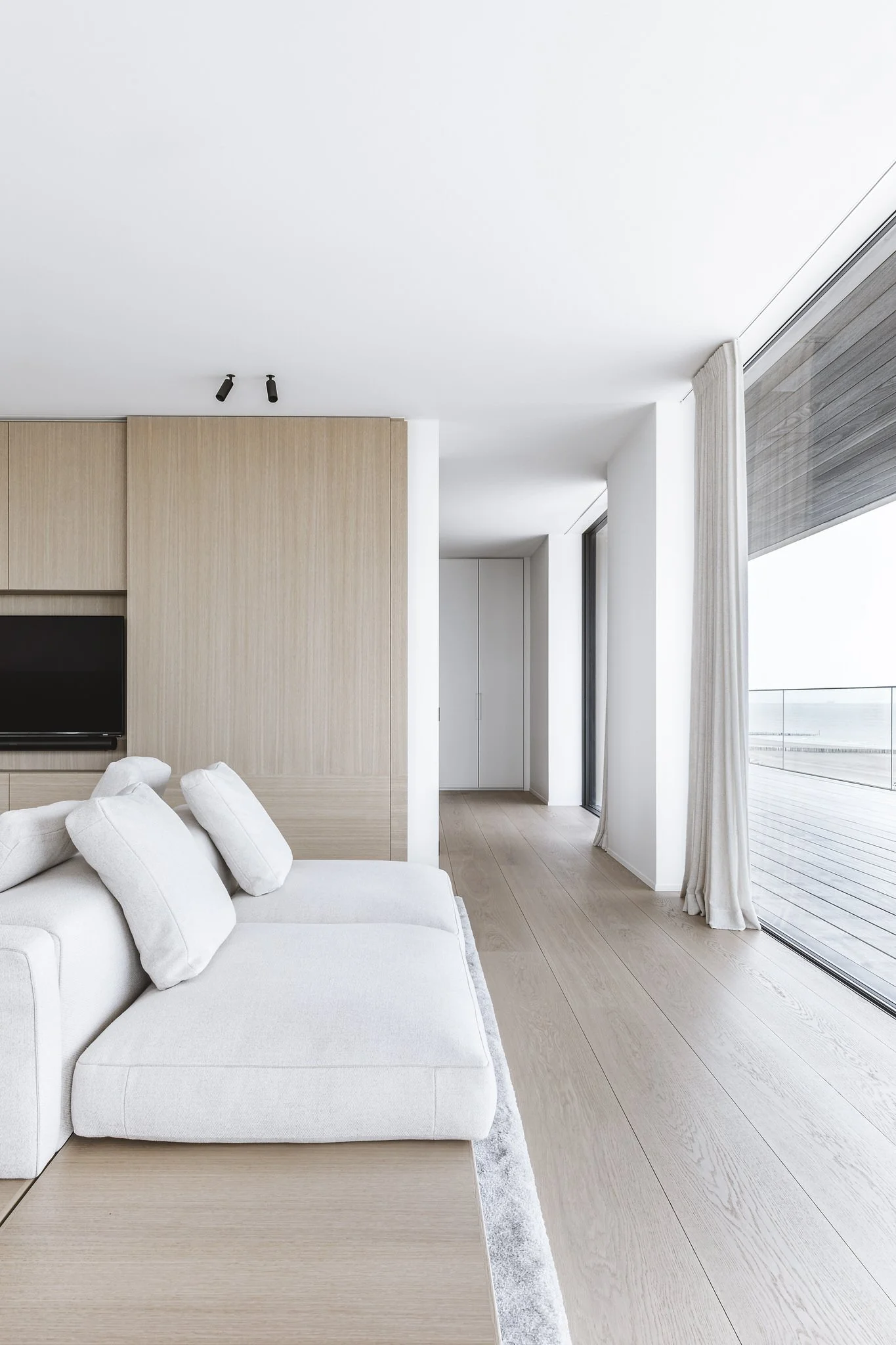 Sala moderna con sofá blanco, madera clara y grandes ventanas con cortinas y vista al exterior