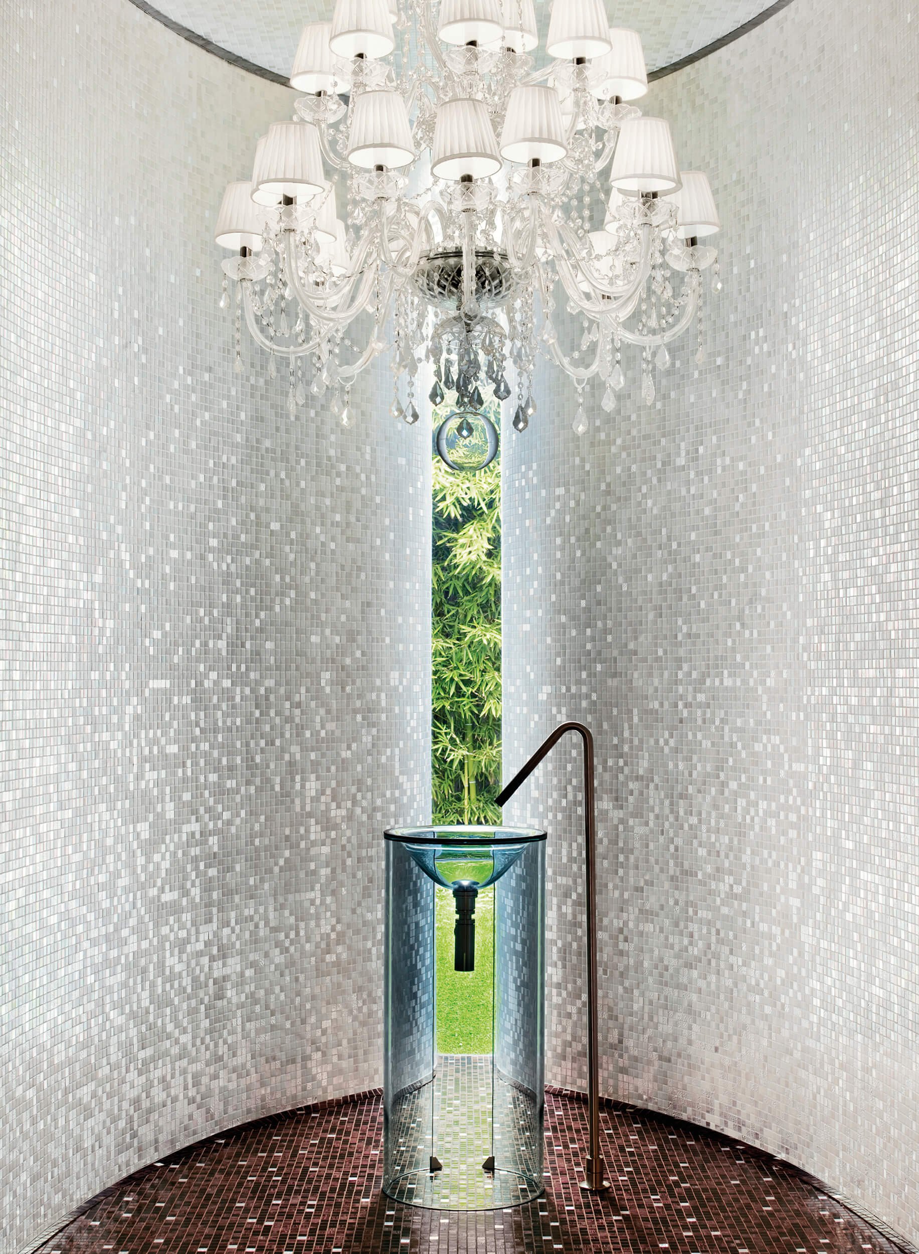 Fuente moderna en un espacio con paredes de azulejos blancos, una lámpara de araña y vista a un jardín con plantas verdes.