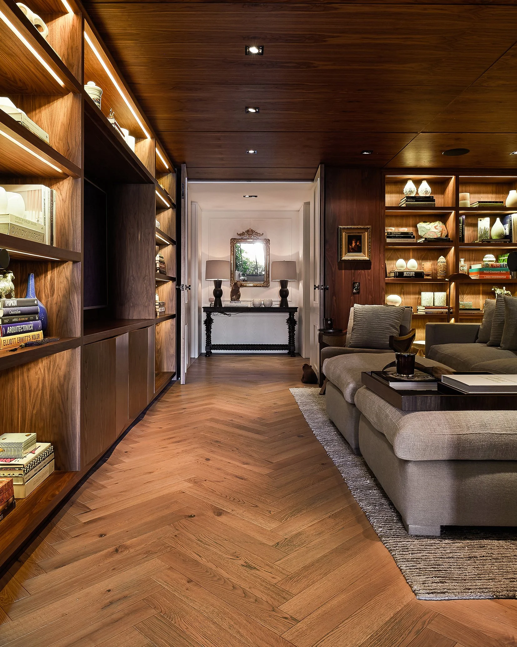 Sala de estar con paredes de madera y estanterías llenas de libros y objetos decorativos, sofá beige con cojines, mesa de centro con libros y vaso, alfombra y una consola con lámparas y adornos al fondo, en una decoración moderna y cálida.